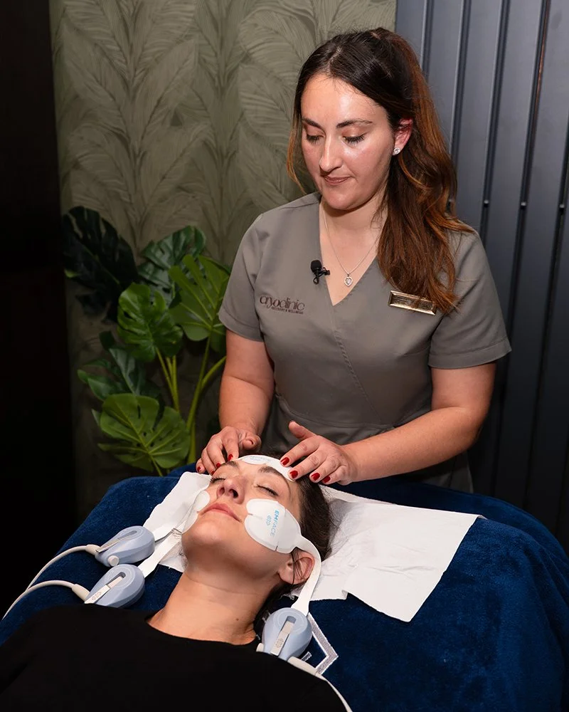 A woman lying on a treatment bed with eye patches and electrodes on her face, receiving an EMface treatment from a skincare professional in a spa or clinic setting.