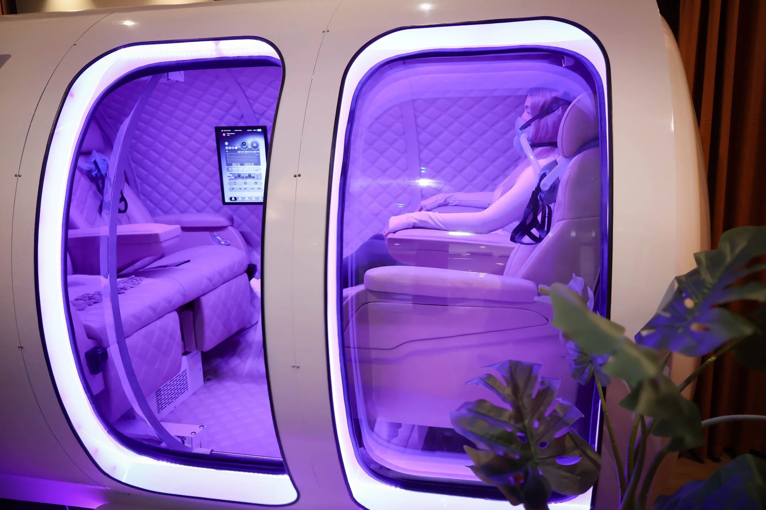 Inside an HBOT capsule with padded quilted walls and cushioned seats, illuminated with purple lighting, and featuring a touchscreen interface on the wall.