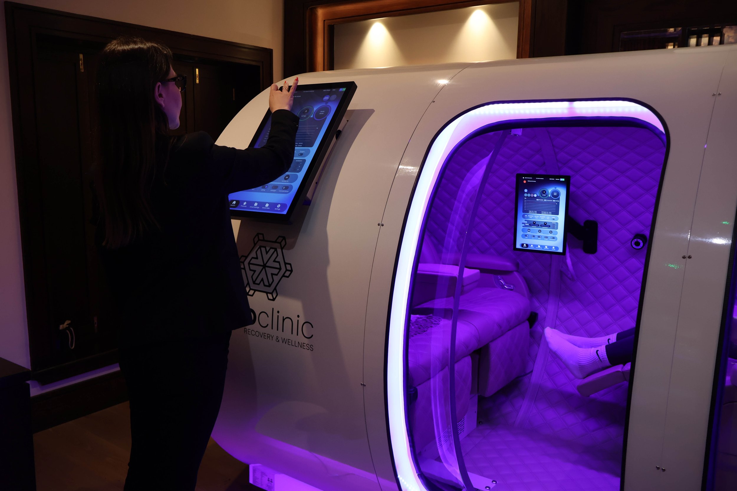 A woman in black clothing and glasses interacts with a touchscreen panel outside a futuristic capsule or pod with purple lighting, which appears to be in a wellness or recovery kiosk.