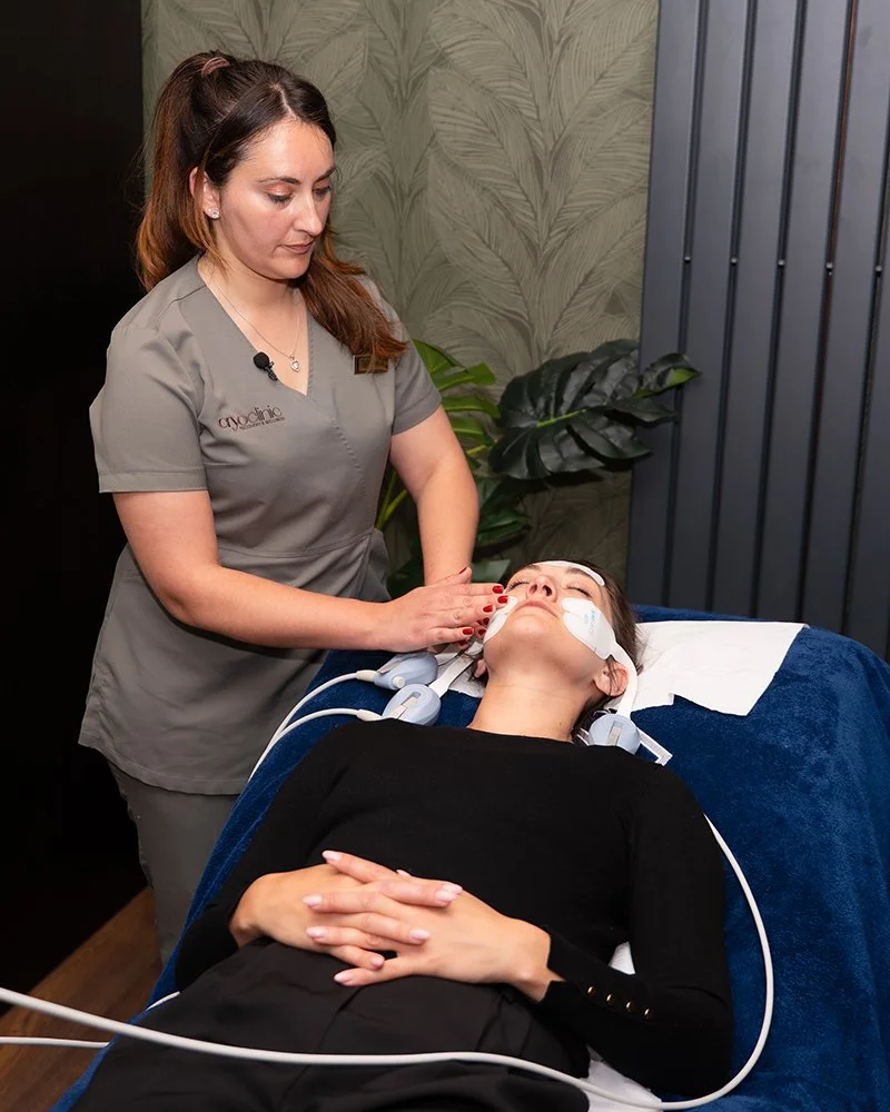 A woman lying on a treatment bed with electrodes on her face and head, receiving an EMFace treatment from a technician in a spa or medical setting.