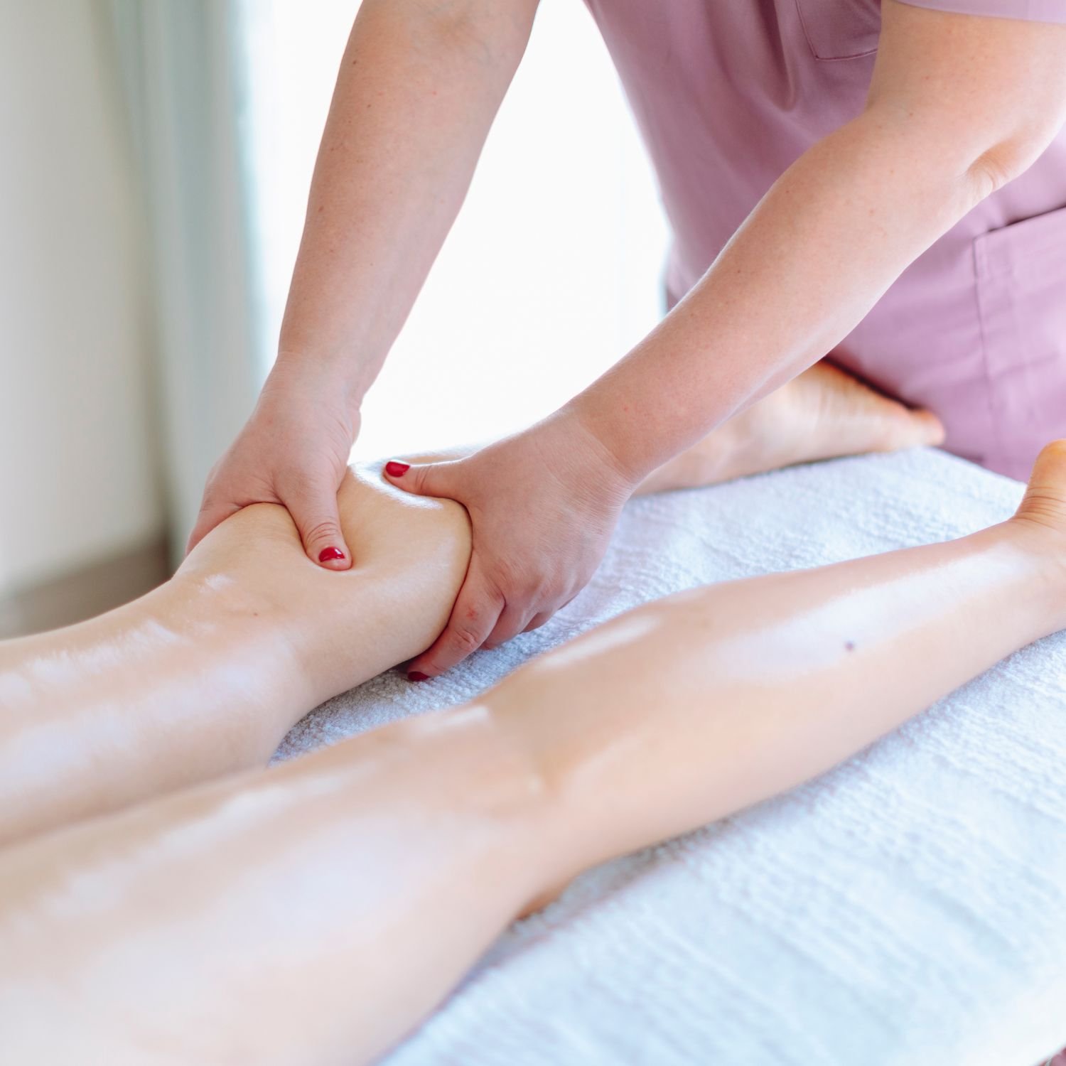 A massage therapist applying massage oil or lotion to a person's leg while lying face down on a massage table.