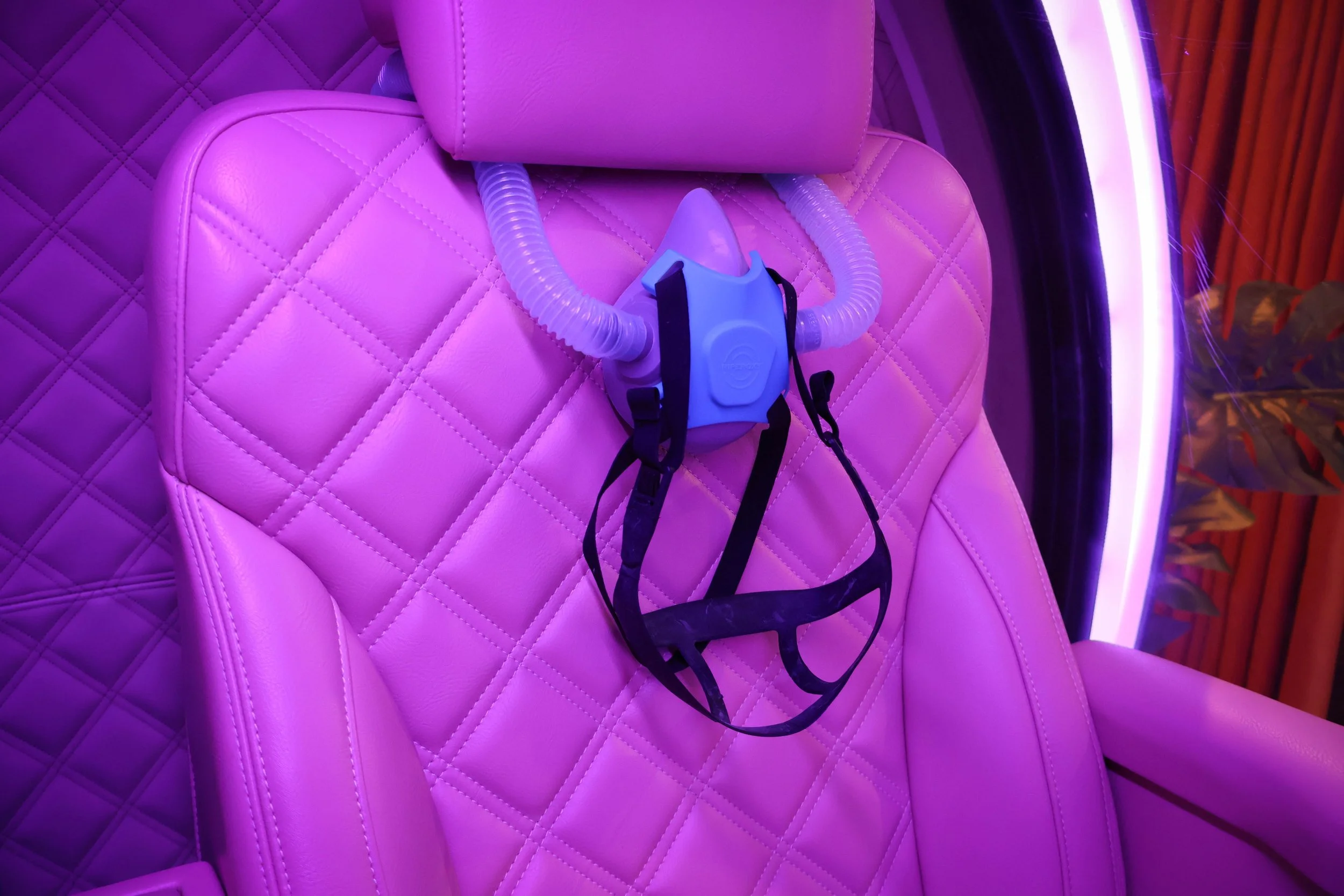 Pink quilted cushioned seat with a breathing mask and straps placed on it, illuminated with purple and pink lighting.