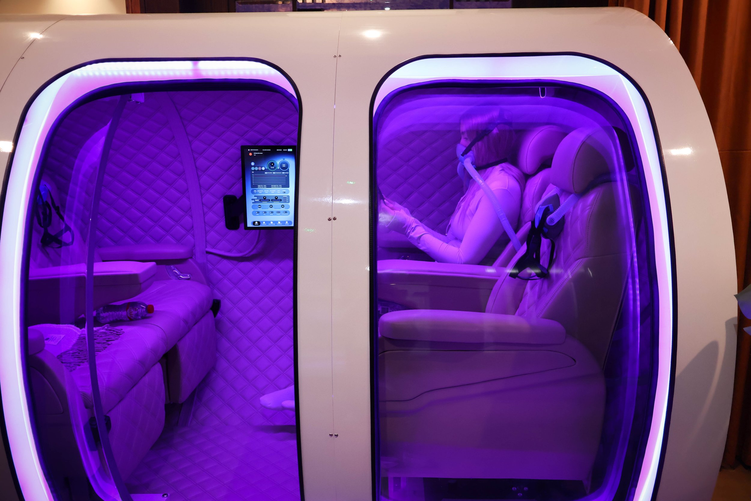 A person wearing a protective suit, gloves, and a mask is inside a futuristic, pod-like structure with purple lighting. The pod has a quilted interior with seating and control panel, suggesting a high-tech relaxation or therapy chamber.