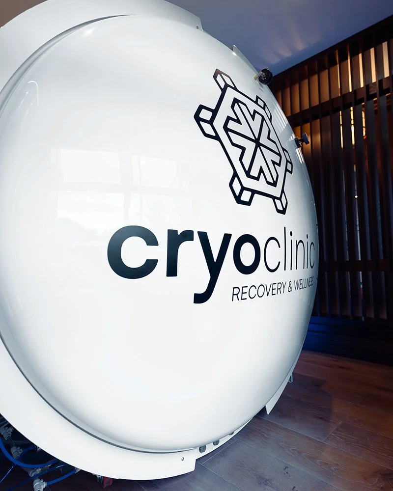 Interior of Cryo Clinic, a large white HBOT tank with the clinic's logo and name visible on its side, next to wooden wall paneling.