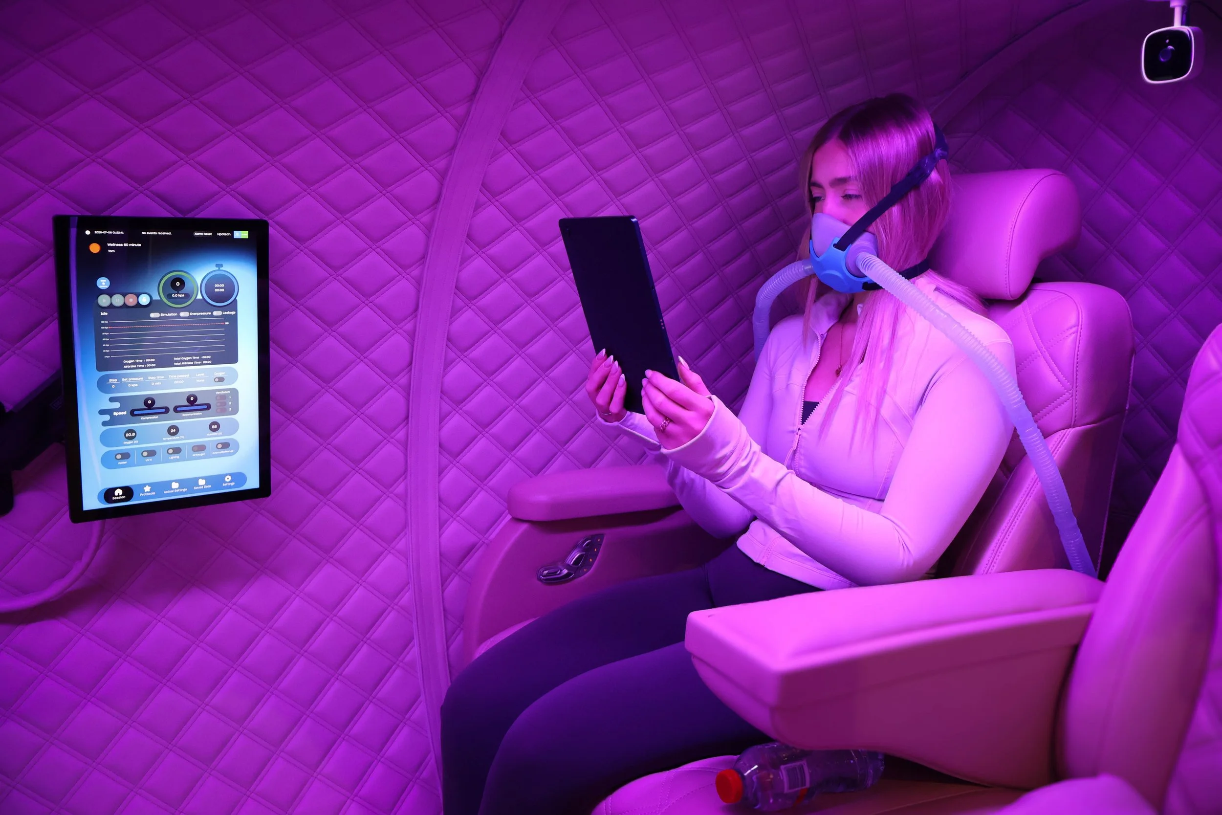A woman is sitting in a cushioned, pink-themed room wearing a mask and breathing apparatus connected to a tube. She is holding a tablet, and there is a screen displaying medical data nearby. The room has a quilted pink wall and a mounted device on th