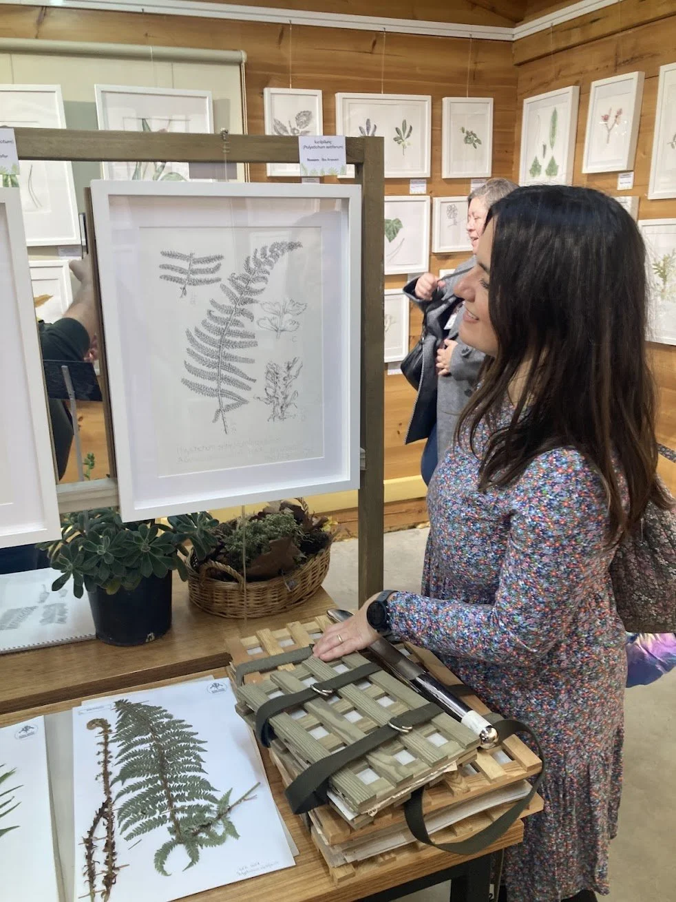 People viewing framed botanical illustrations of ferns and plants at an art or botanical exhibit.