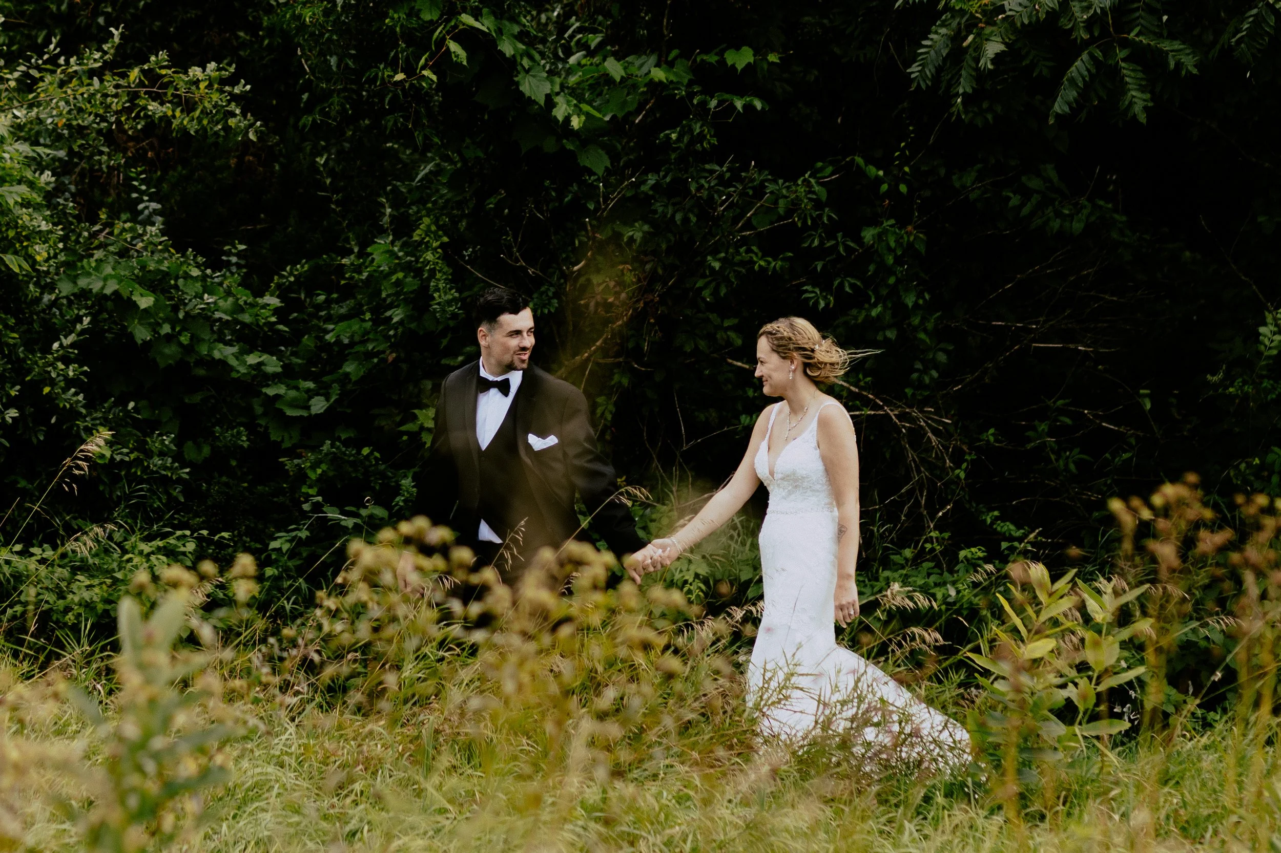 Newlywed couple holding hands and walking through a lush green field, with the groom in a black tuxedo and the bride in a white wedding dress.