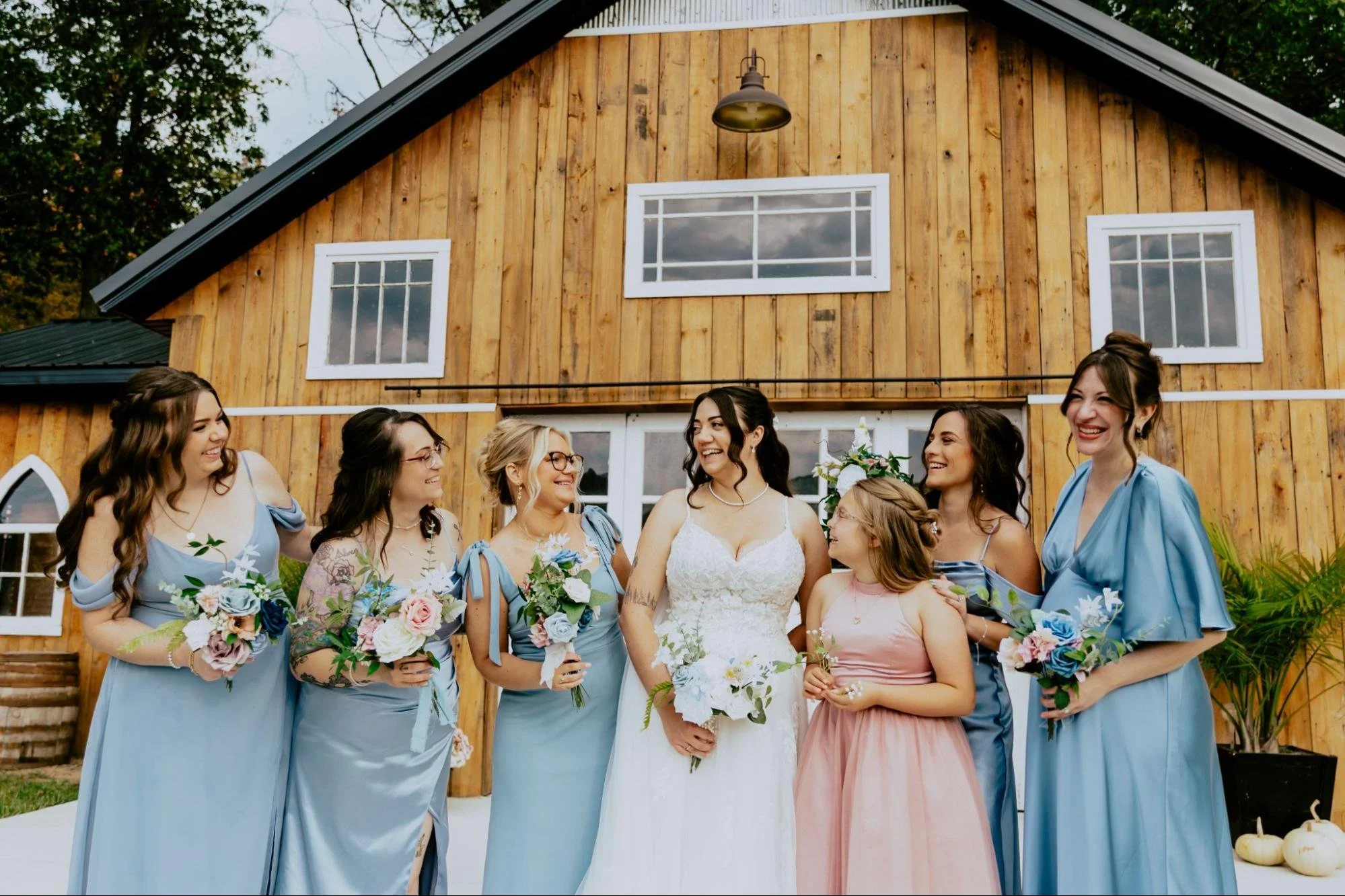 Detroit wedding photographer capturing a joyful bride with her bridesmaids and flower girl in front of a rustic barn wedding venue.