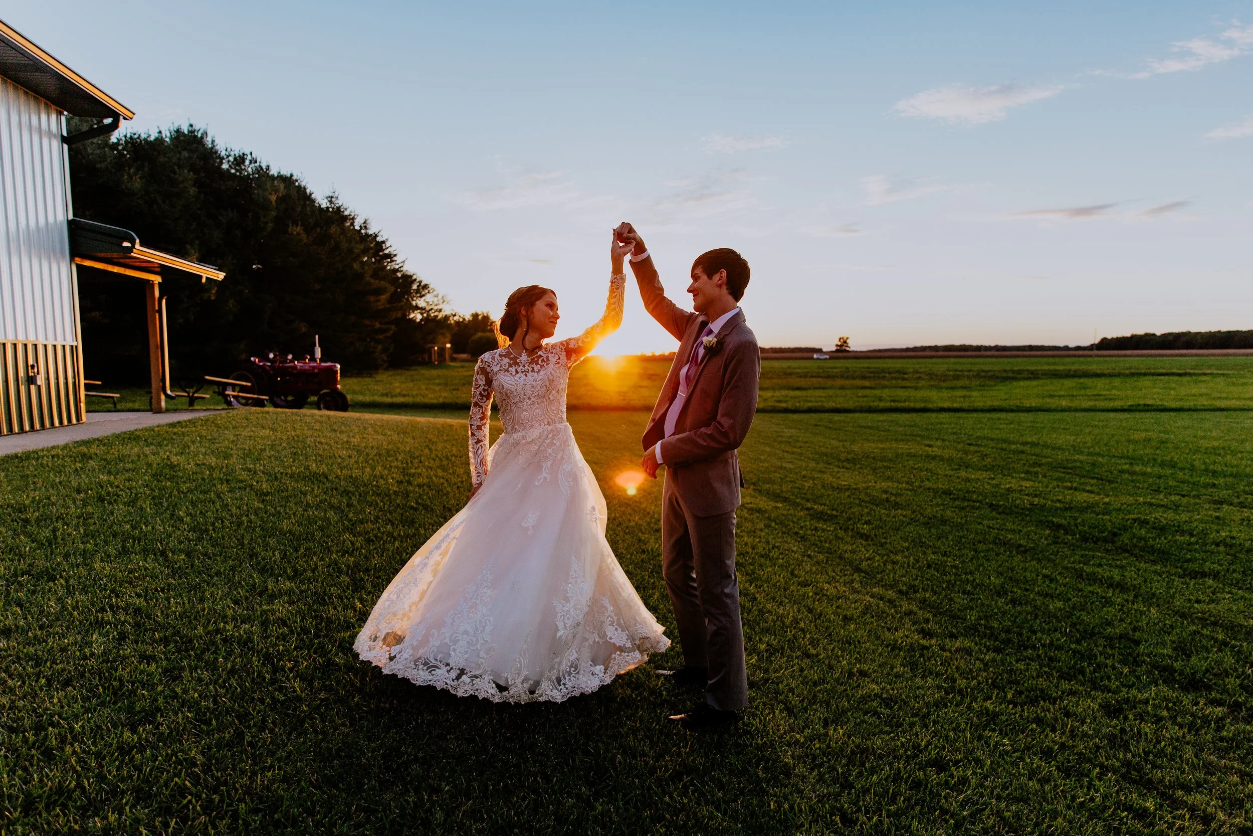 Newlywed couple dancing together in a grassy field at sunset, with the bride in a lace wedding dress and the groom in a suit as the sun sets behind them.