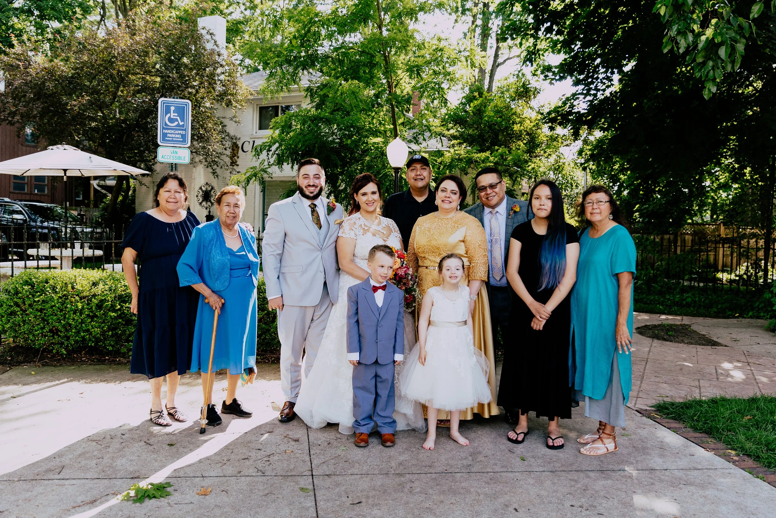 Newlywed couple posing with their family and children outside their wedding venue, surrounded by trees and greenery.