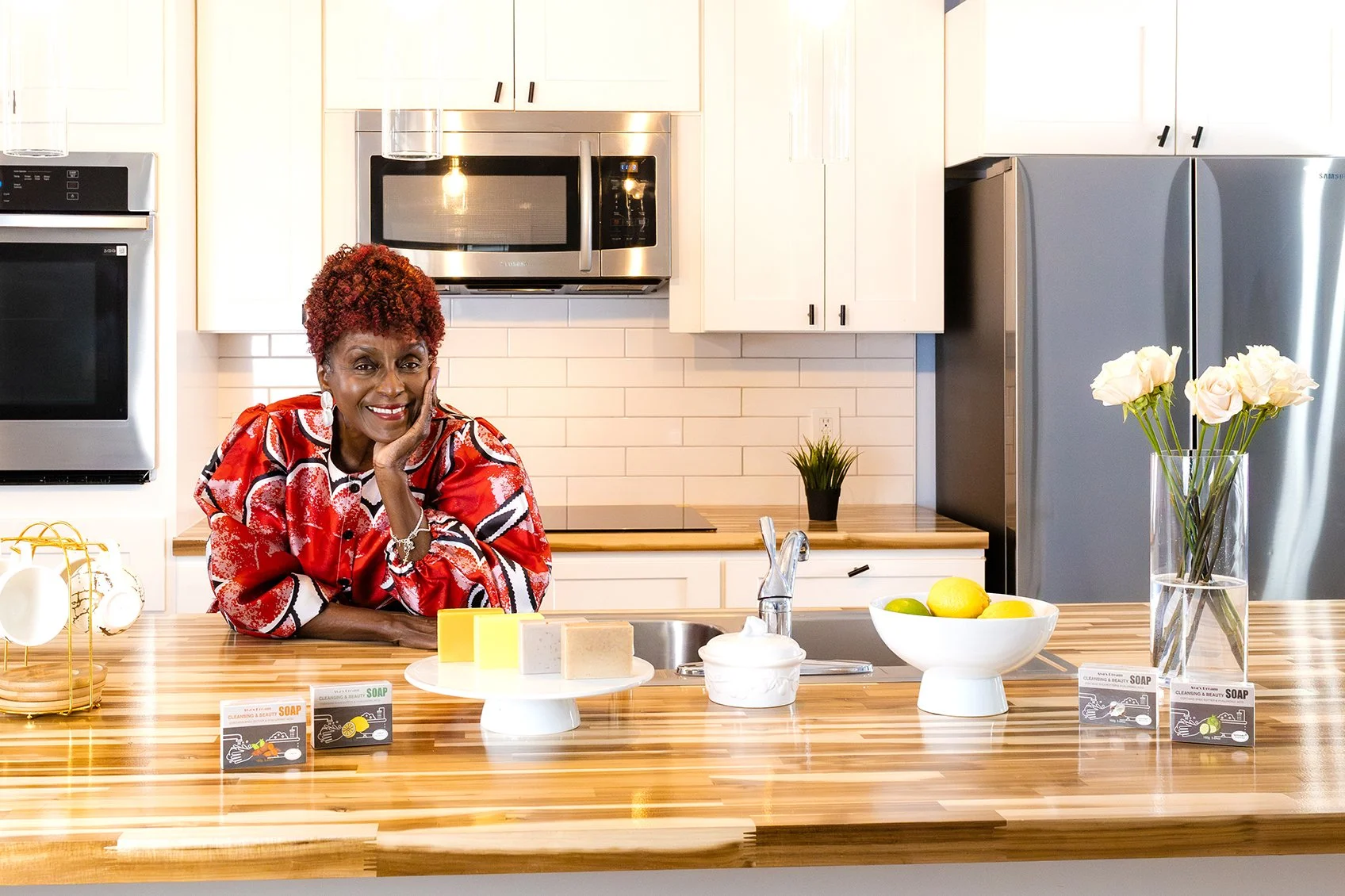 Detroit Entrepreneur Ava Jackson standing in a kitchen at the counter leaning looking at the camera smiling with her soap in front of her.