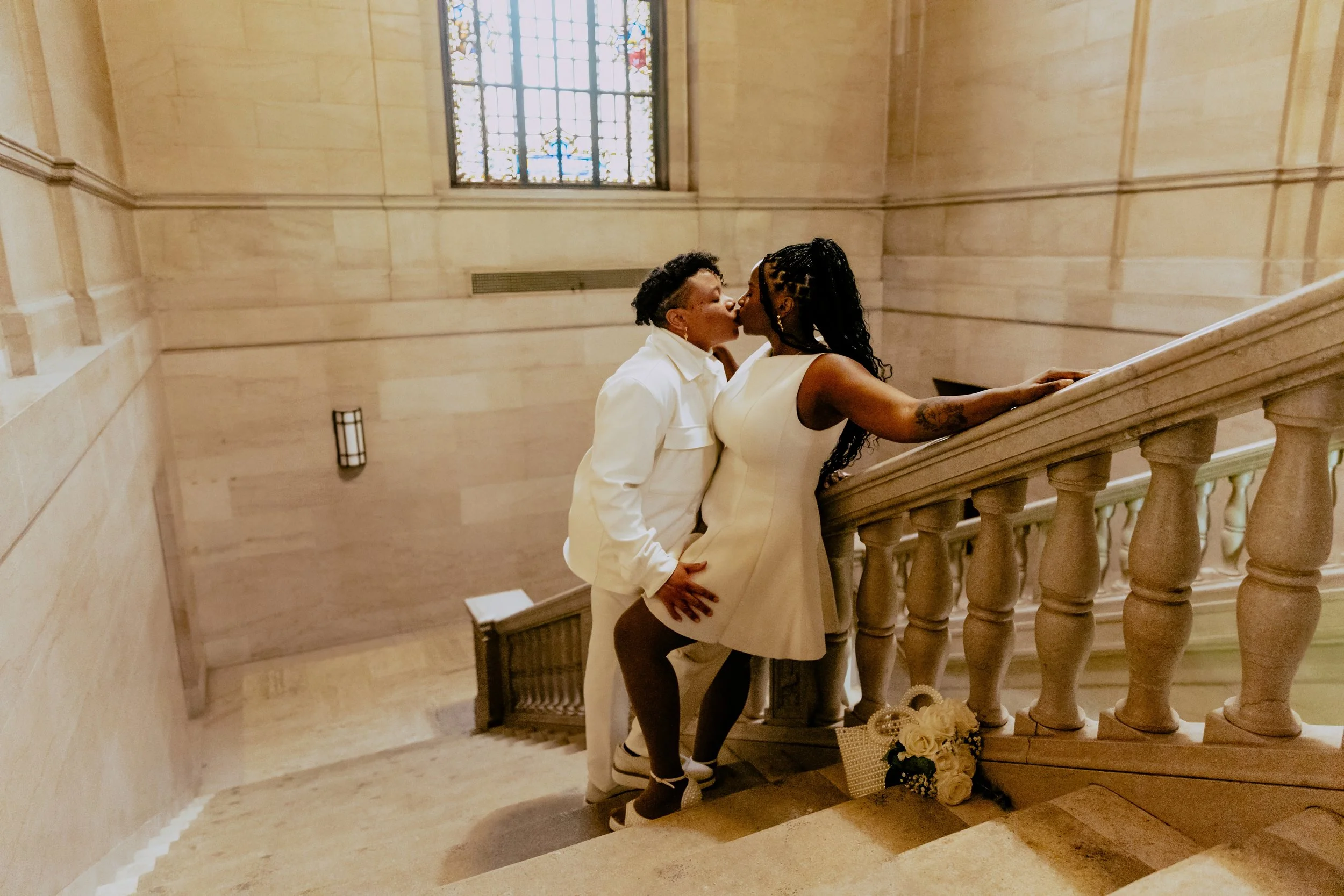 Two brides share a kiss on a grand staircase inside an elegant wedding venue, with their bouquet resting on the steps.