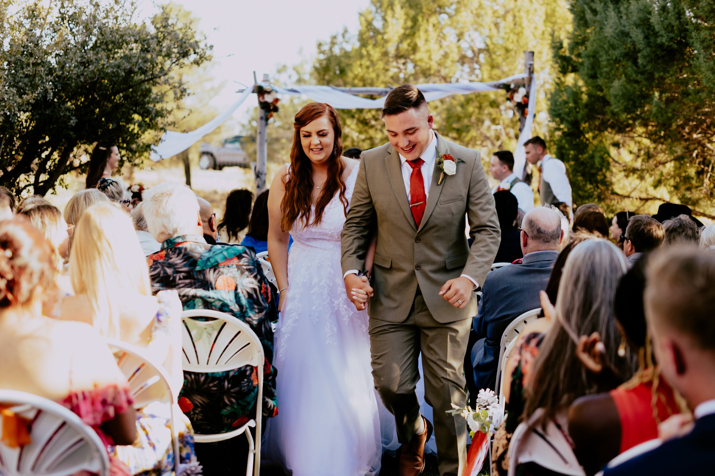Newlywed couple walking hand in hand down the aisle after their outdoor wedding ceremony, surrounded by smiling guests and greenery.
