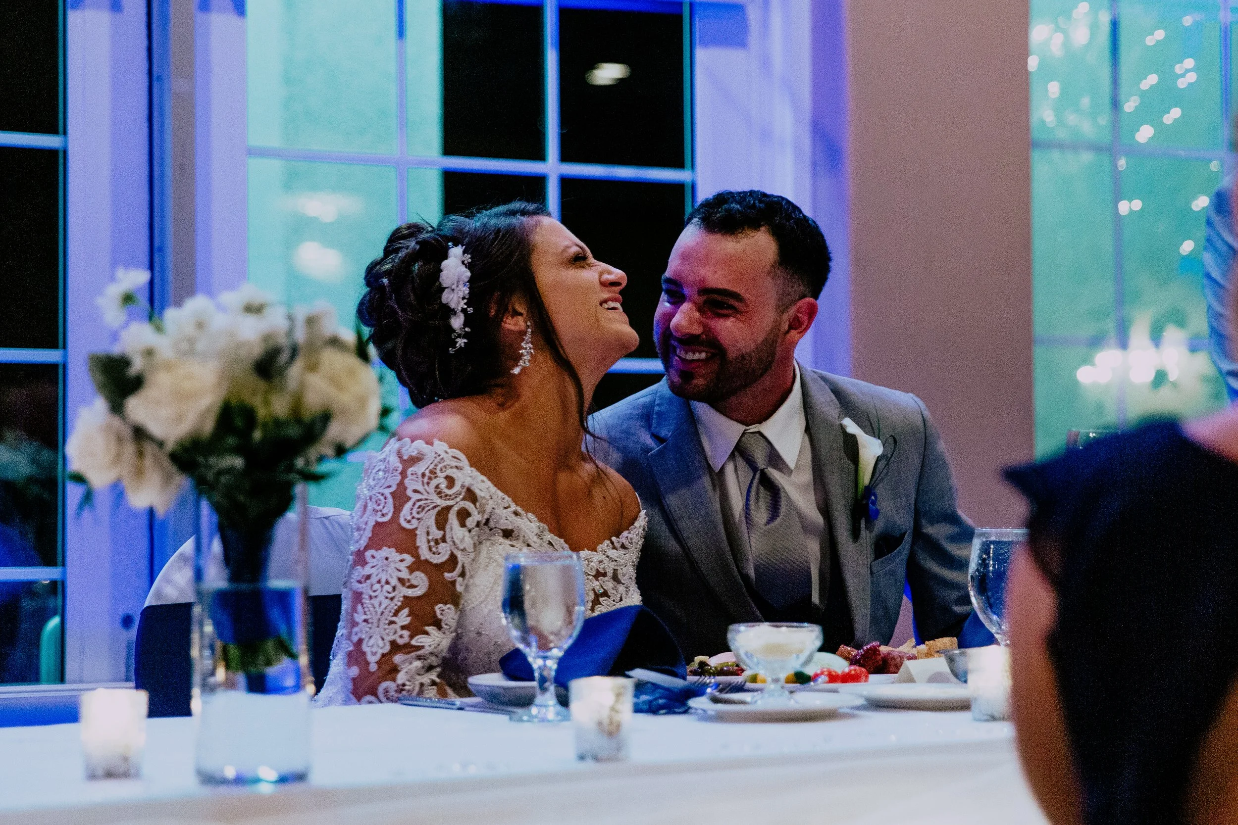 Newlywed couple laughing and sharing a joyful moment together at their wedding reception while seated at the dinner table.