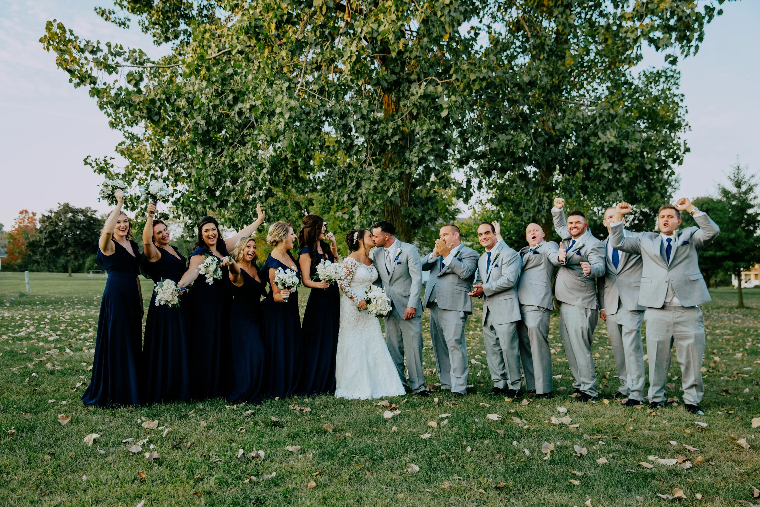 Bride and groom pose with their wedding party outdoors under a large tree, with bridesmaids in navy dresses and groomsmen in gray suits celebrating around them.