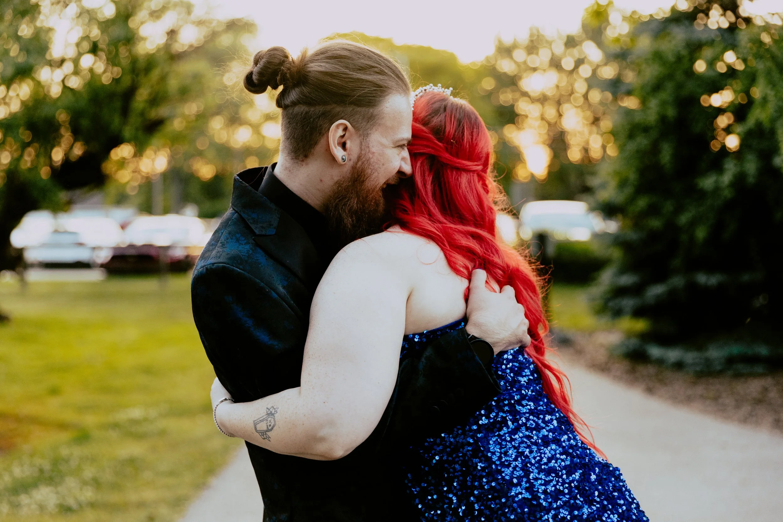 Couple embracing outdoors at golden hour, with one partner wearing a black suit and the other wearing a sparkling blue dress with long red hair.