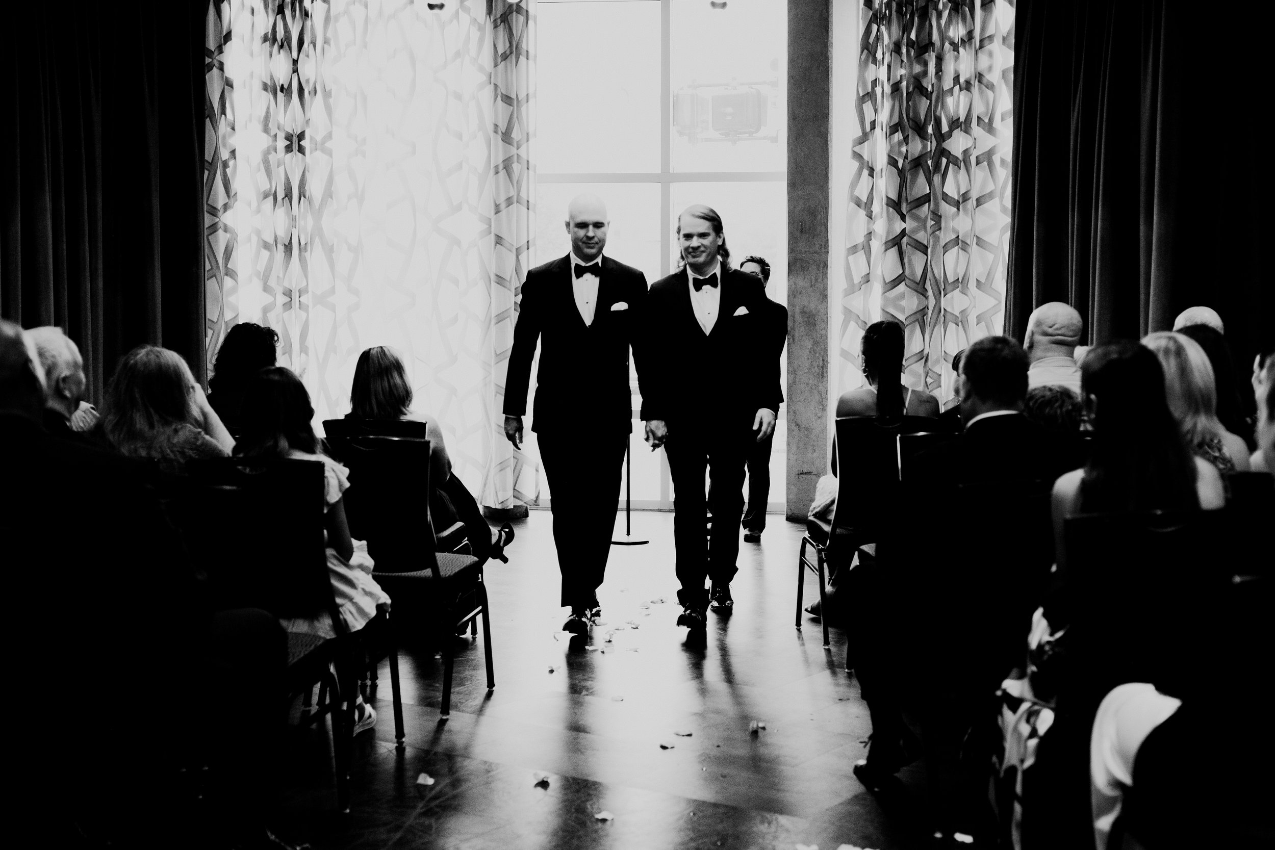 Two grooms in black tuxedos walk hand in hand down the aisle during their wedding ceremony as guests watch from their seats.