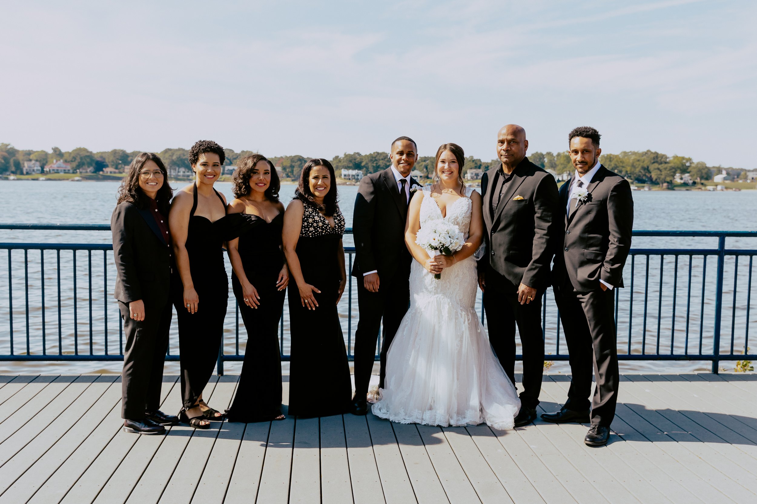 Wedding couple posing with their wedding party on a waterfront deck, with the bride holding a white bouquet and the group dressed in formal attire.