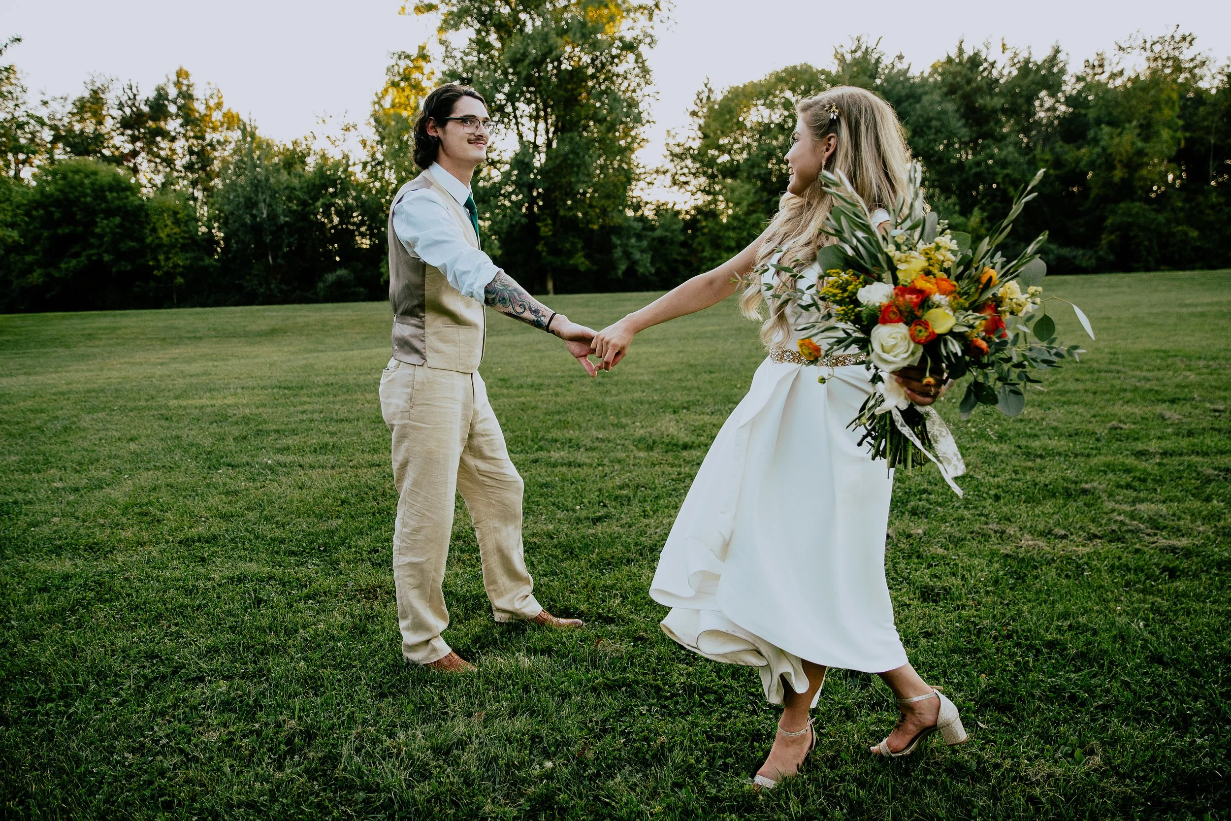 Newlywed couple holding hands and walking together across a grassy field, with the bride carrying a colorful bouquet.