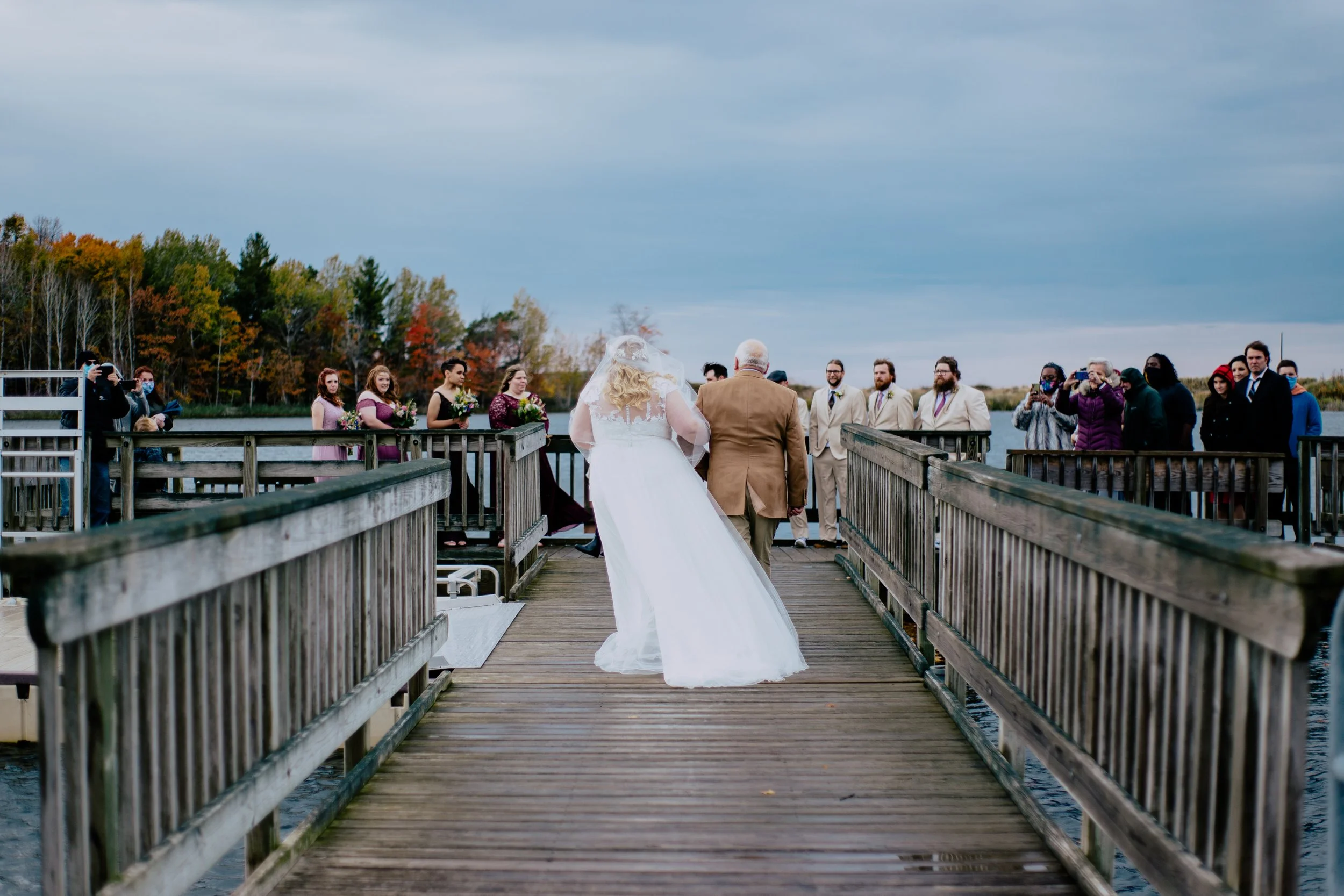 Bride and dad walking down a wooden lakeside pier toward wedding guests during an autumn wedding ceremony.