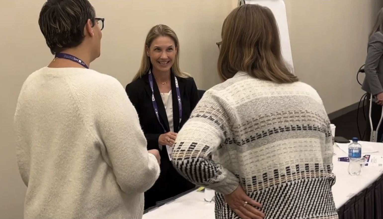 Three women are engaged in a conversation at a conference or meeting. One woman, wearing a black blazer, is smiling and talking to two women with their backs facing the camera. The women with their backs to the camera are wearing sweaters, and there are water bottles and papers on the table in front of them.