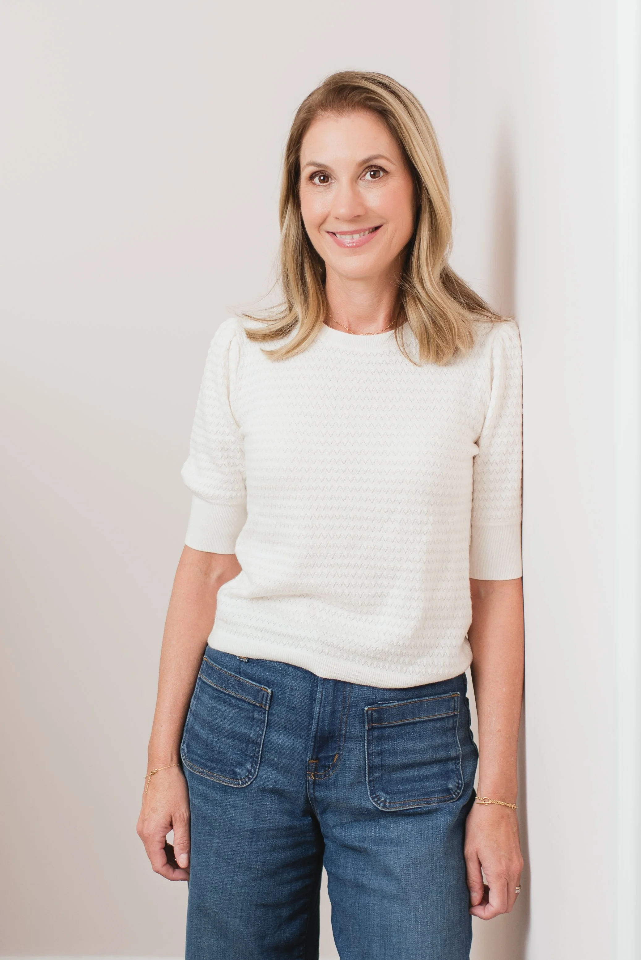 A woman with shoulder-length blonde hair, wearing a white textured short-sleeve sweater and blue jeans, standing against a plain light-colored wall and smiling at the camera.