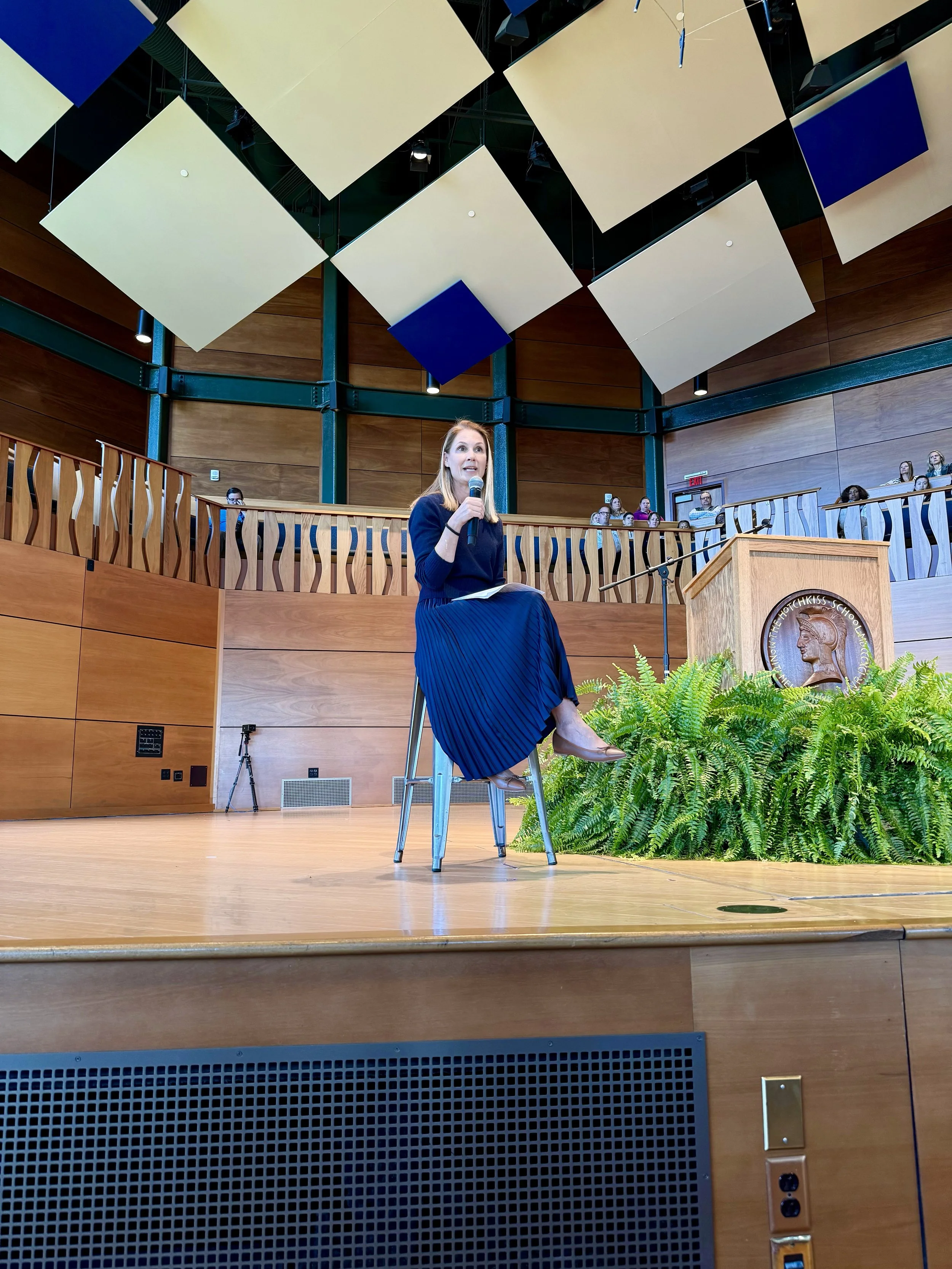 A woman sitting on a high stool with a microphone, speaking to an audience in an auditorium or lecture hall. She is wearing a dark top and a long, pleated skirt. There are people watching in the background, some wearing masks. The room features wooden panels, a podium with an emblem, lush green plants, and square acoustic panels on the ceiling.
