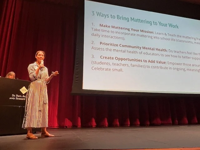 A woman giving a presentation on a stage with a large screen displaying three ways to bring mattering to work. The slide reads: 1. Make Mattering Your Mission, 2. Prioritize Community Mental Health, 3. Create Opportunities to Add Value. The woman is wearing a white and black vertically striped dress and holding a microphone. There is a podium with a sign that says 'St. Paul AL'.