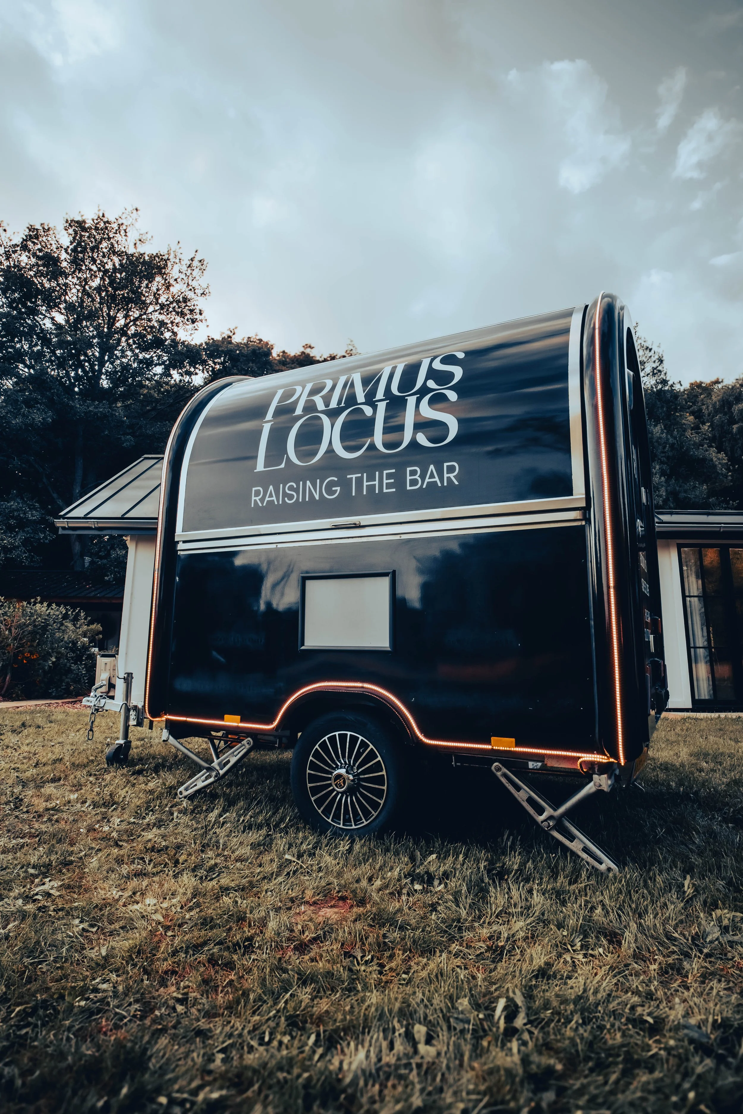 A black food truck with the words "Primus Locus" and "Raising the Bar" written on it, parked on a grassy lawn with trees and a house in the background.