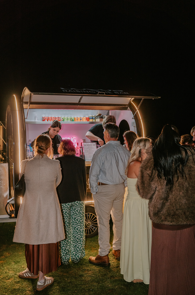 People standing in line at a nighttime food truck or mobile bar, with a bartender serving drinks inside.