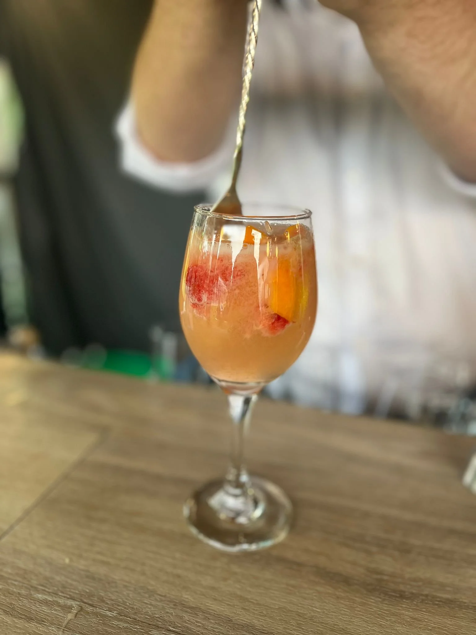 A person pours a drink with fruit pieces into a tall wine glass on a wooden surface, with a blurred background.