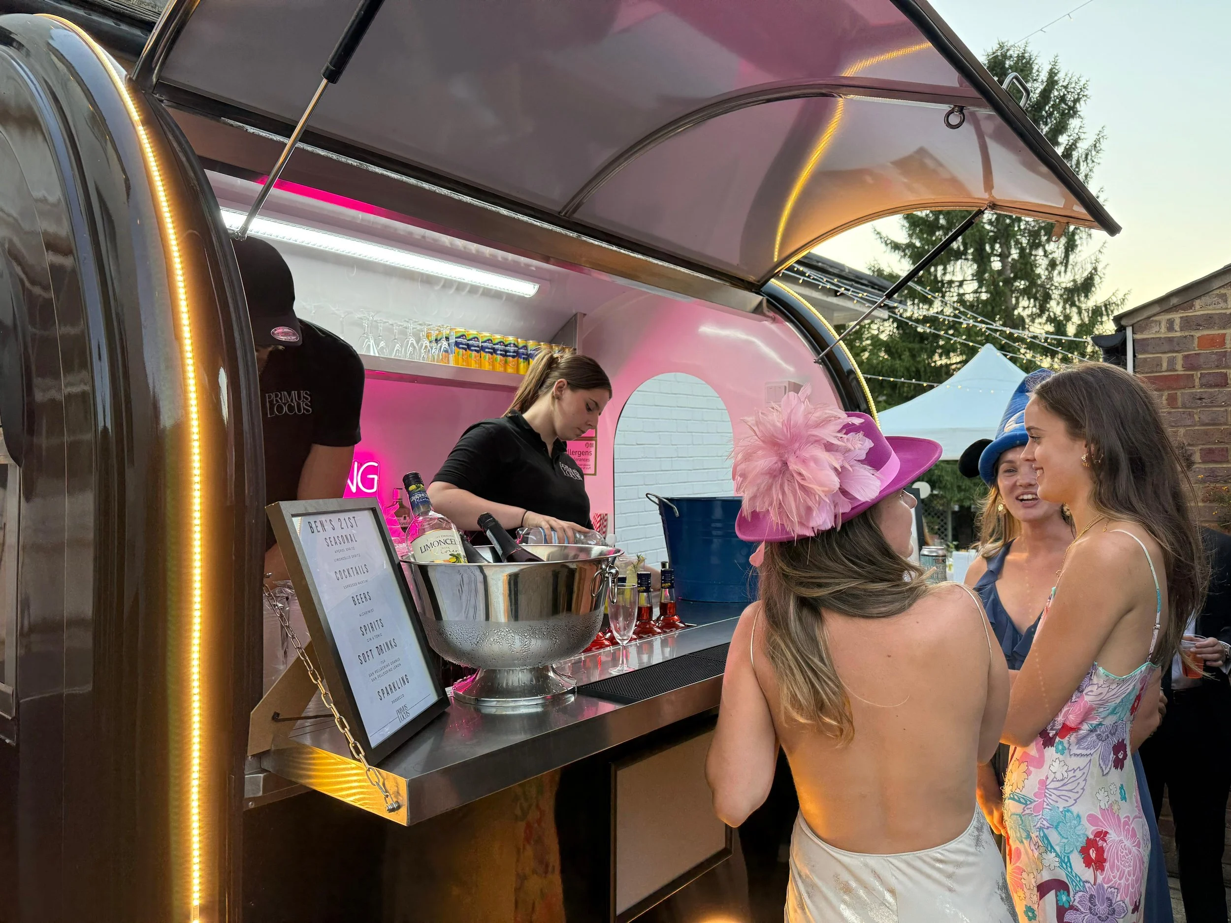 People dressed in colorful outfits and hats socializing near a vintage-style pink and metallic bar trailer at an outdoor event.