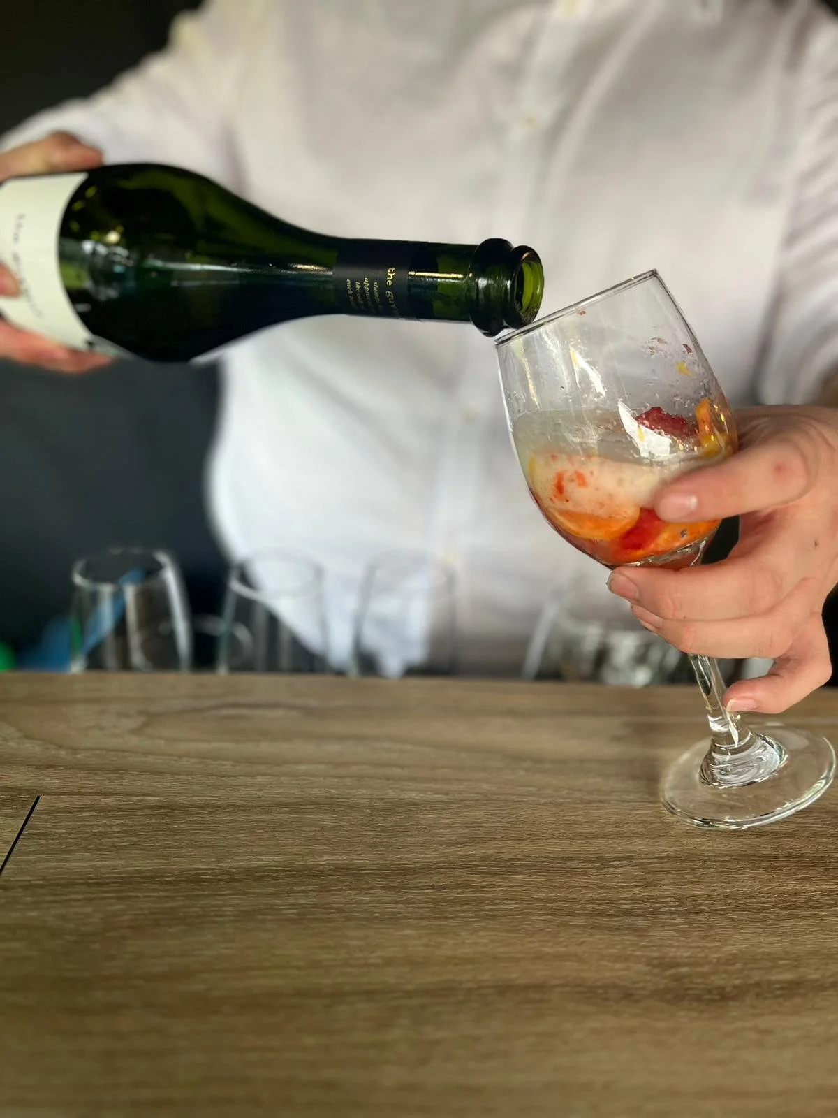 Person pouring a drink from a green bottle into a wine glass with strawberries inside.