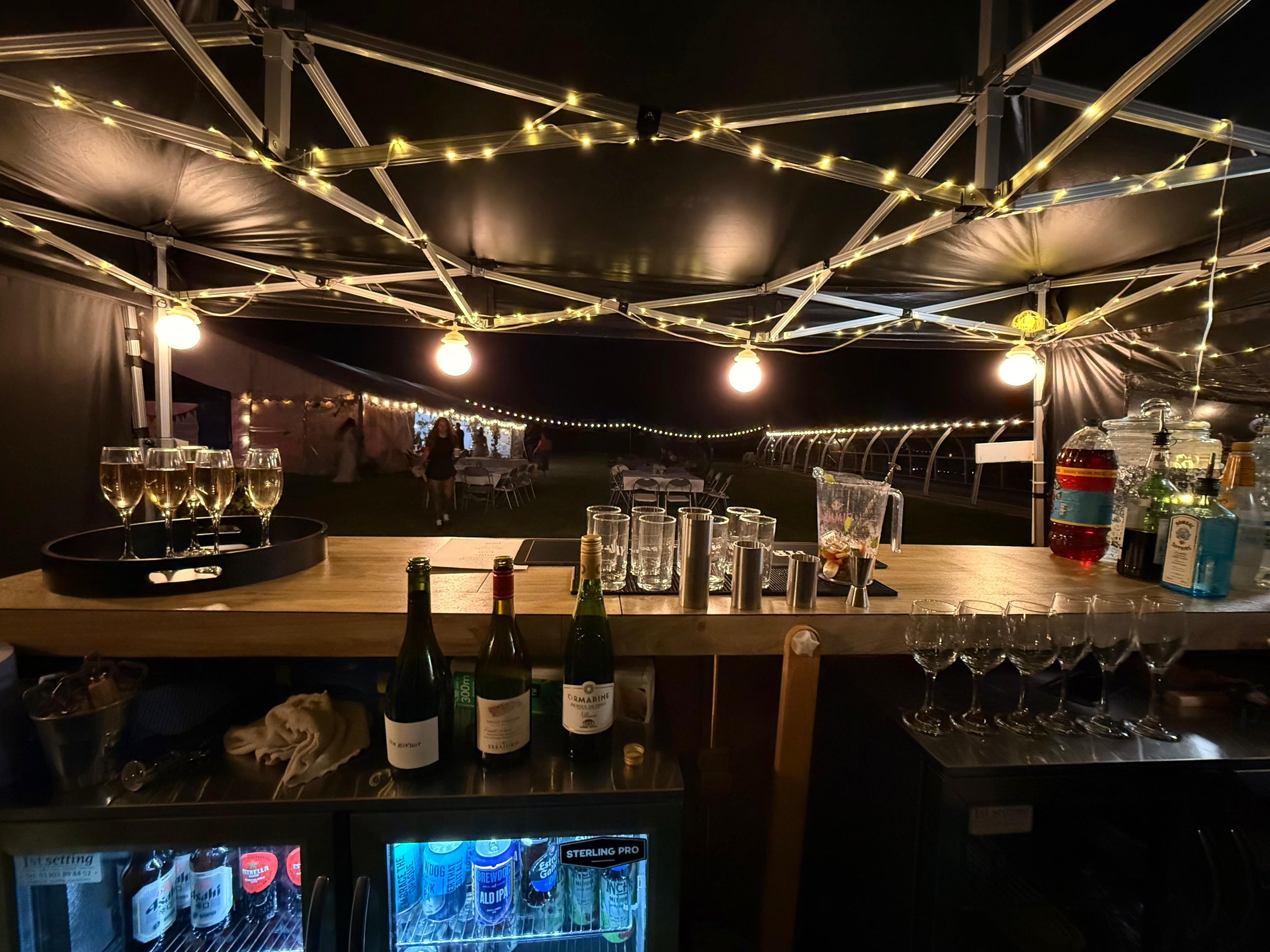 Nighttime outdoor bar under a canopy with string lights and hanging bulbs, with a table and drinks set up inside, several bottles and glasses on the counter, and patio tables with people in the background.