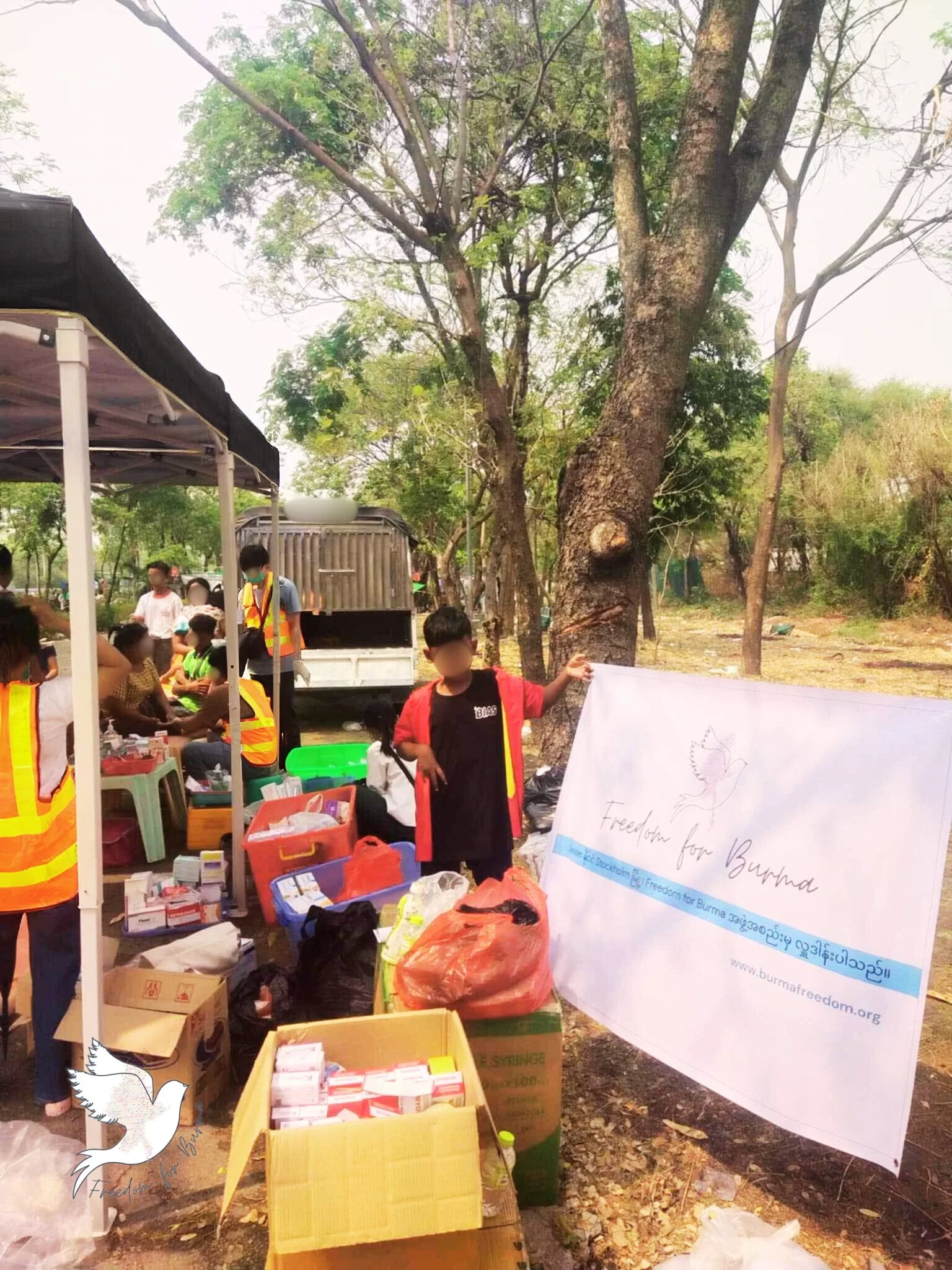 Outdoor charitable event with volunteers distributing supplies, a banner reading 'Freedom for Burma,' and tables with boxes and packages under a canopy in a park-like setting.