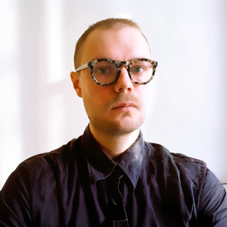 A young man with short hair, wearing large patterned glasses and a black button-up shirt, standing against a plain white background.
