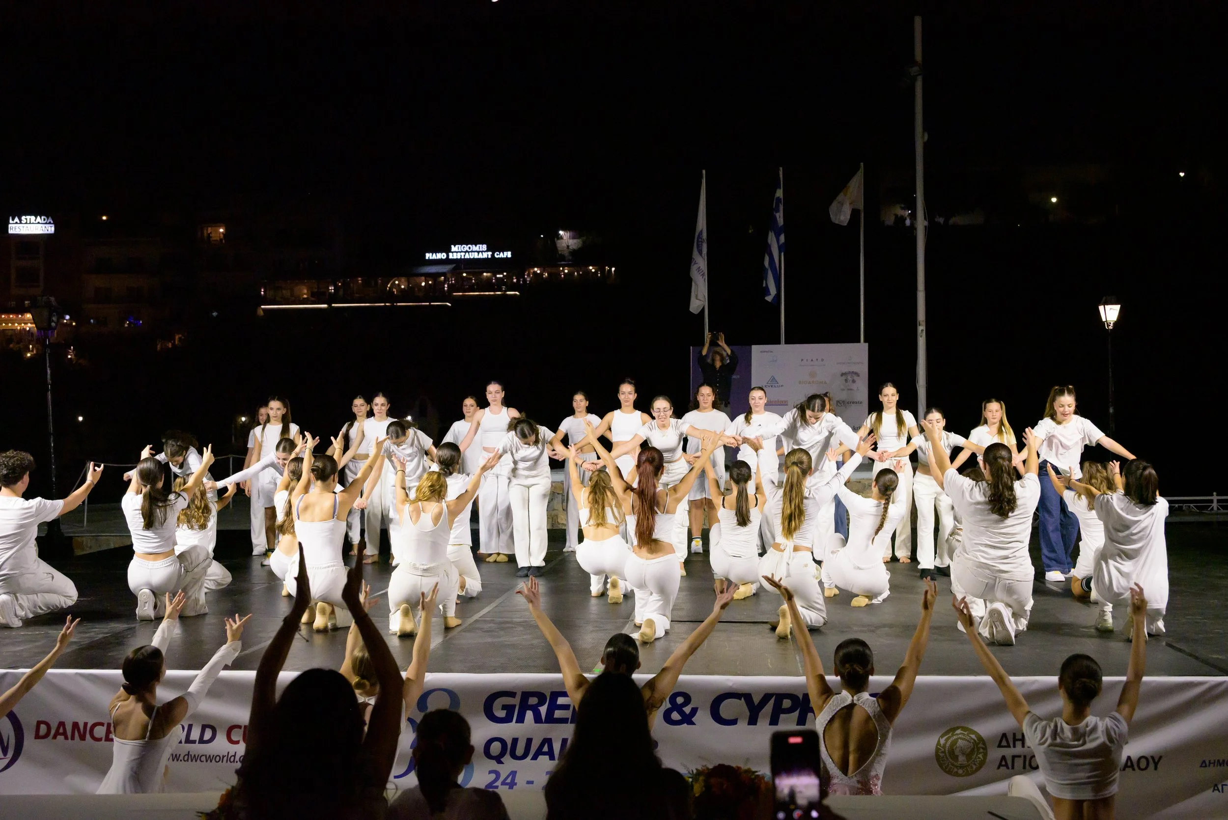 A group of dancers in white outfits performing on an outdoor stage at night, with an audience watching. There are flags and illuminated signs in the background.