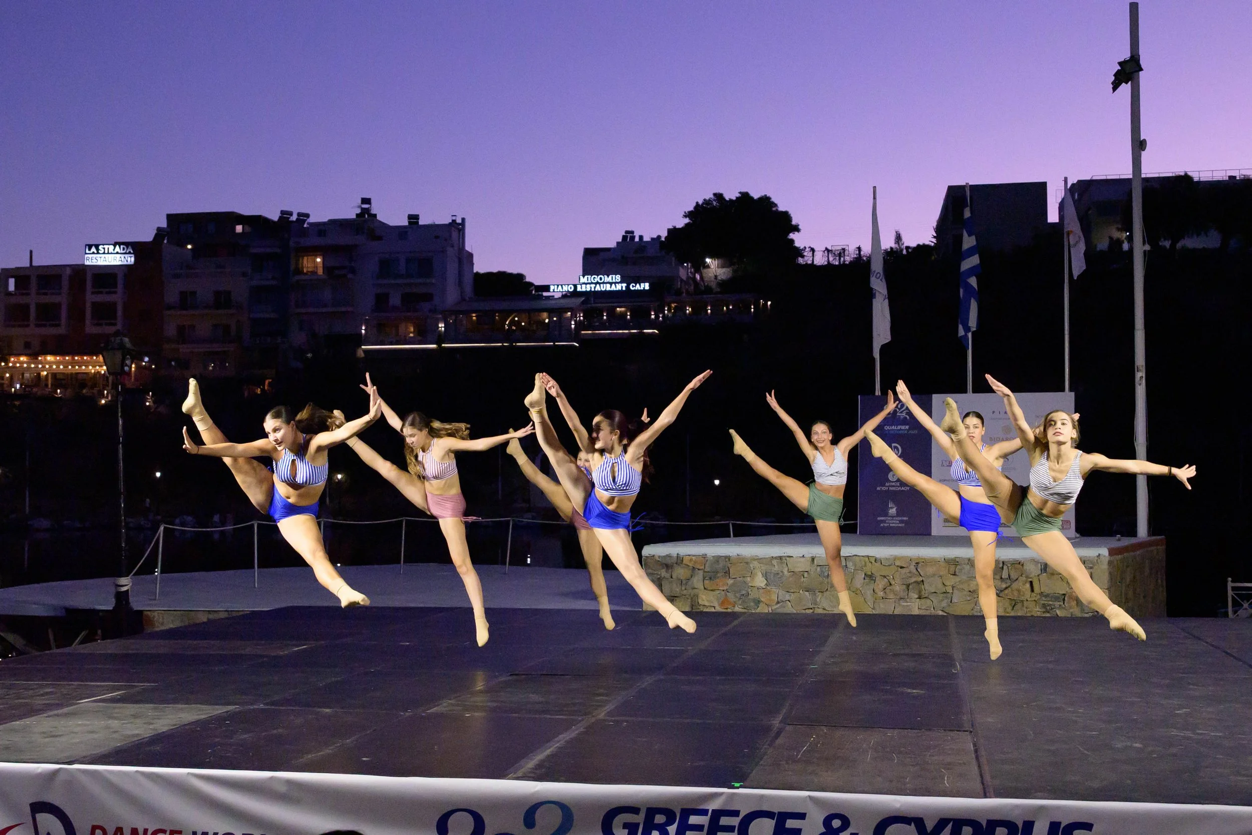 Group of young female dancers performing a synchronized routine on an outdoor stage during dusk, with a purple sky and illuminated buildings in the background.