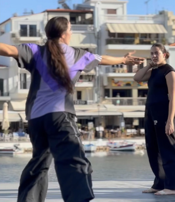 Two women are practicing martial arts or self-defense on a dock near the water, with boats and multi-story buildings in the background.