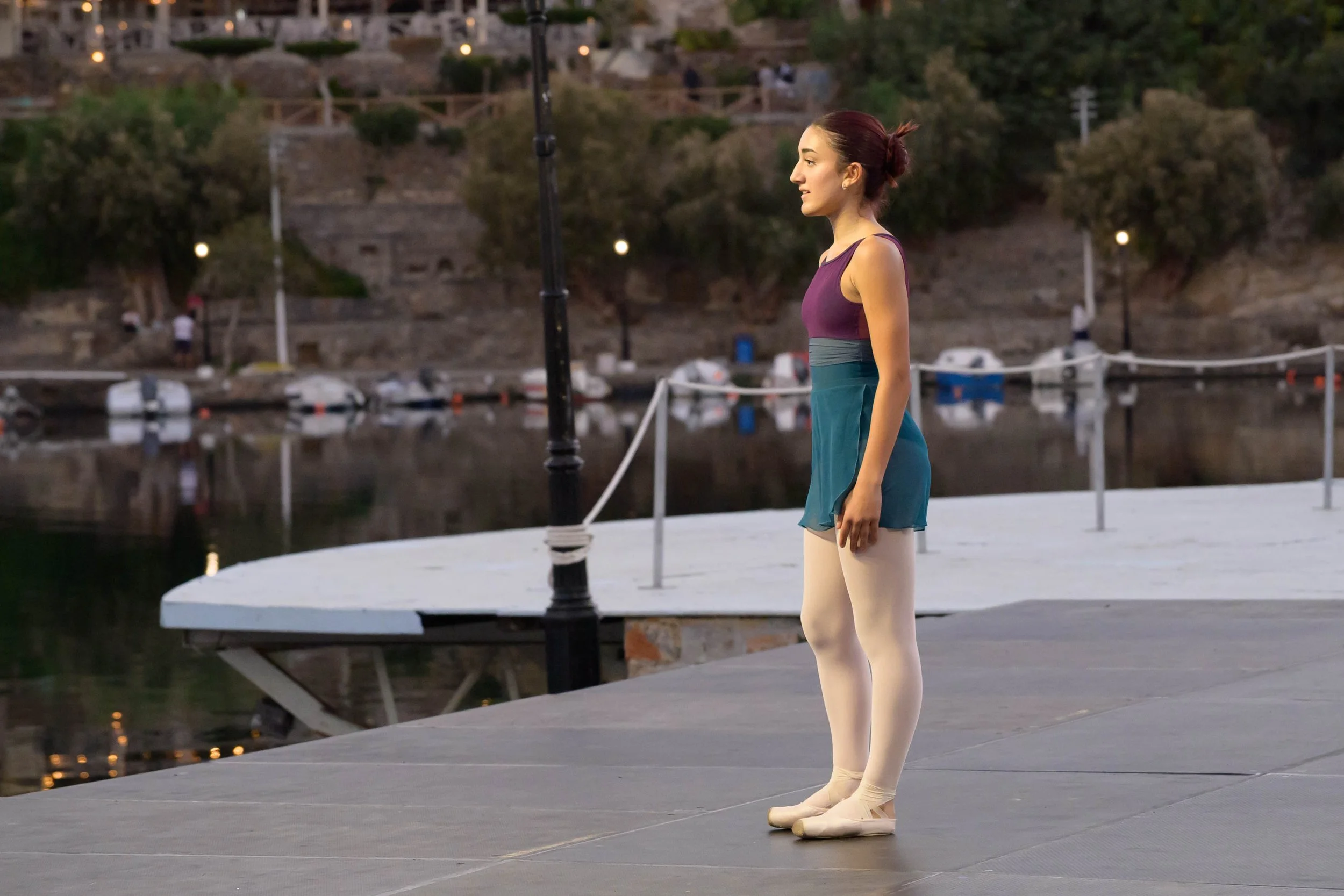 Ballet dancer in ballet slippers standing on a dock by the water during dusk, with a person in the background and boats docked along the water.