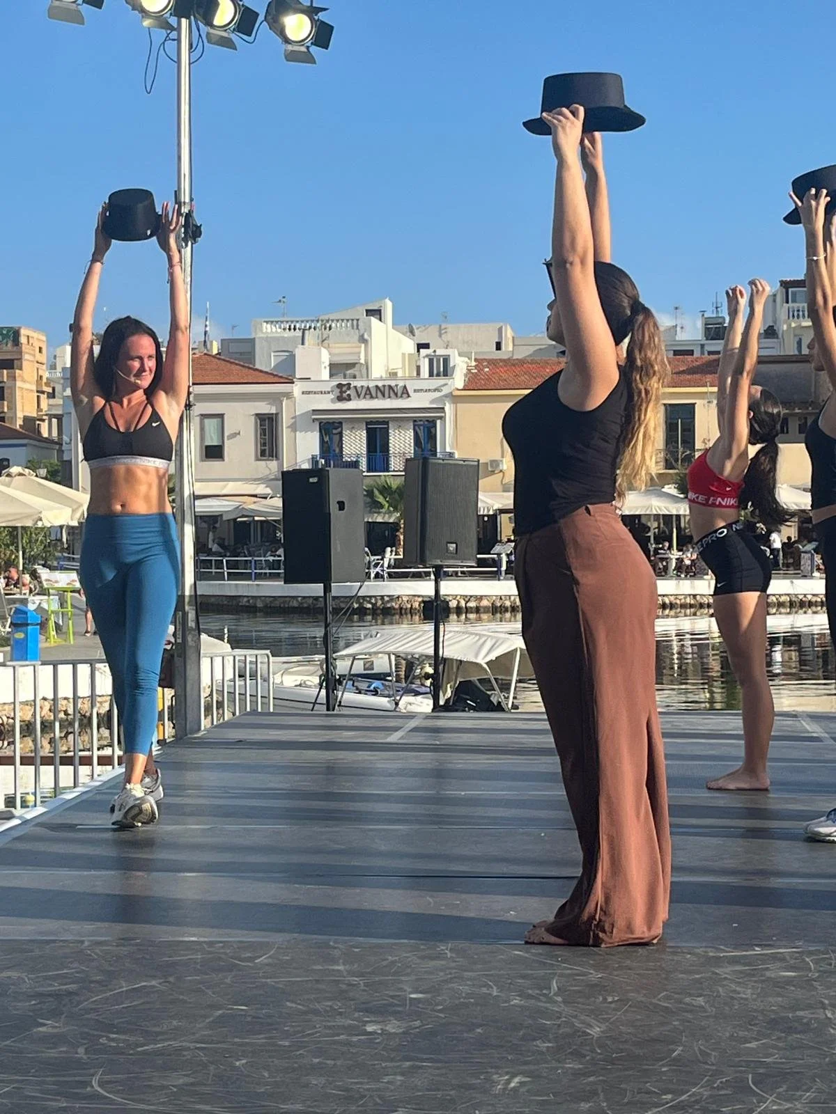 Women practicing yoga or dance on an outdoor stage near water with boats, buildings, and a restaurant in the background.