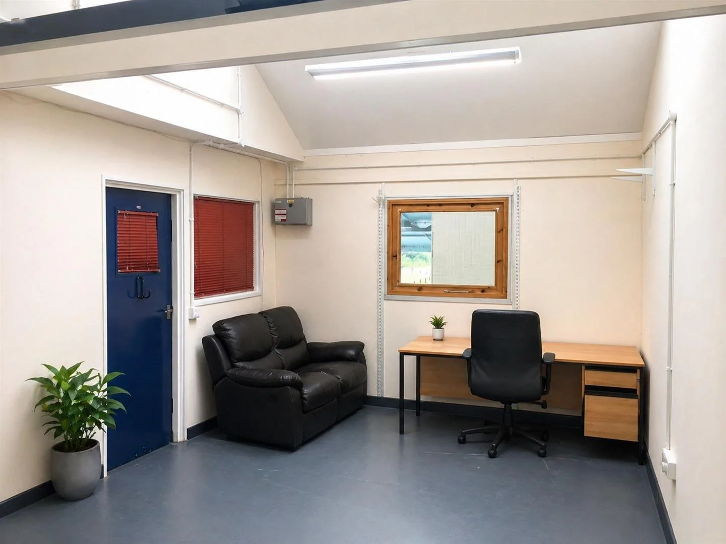 An office room with a black leather couch, a wooden desk with an office chair, a window with wooden framing, two potted plants, red window blinds, a blue door with a red vent, and fluorescent ceiling lights.