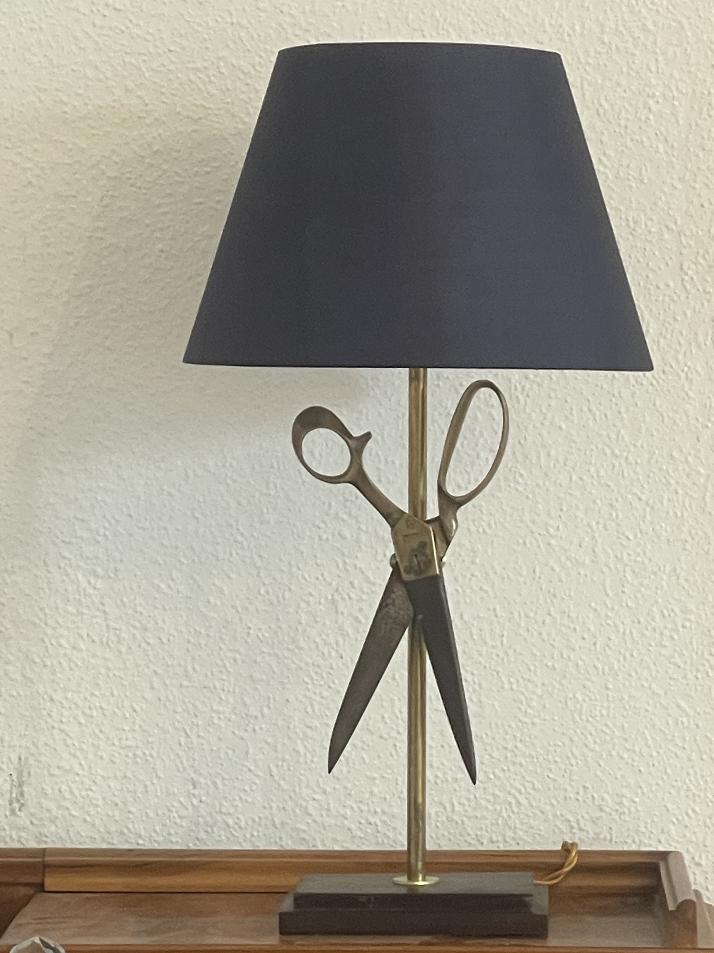 Table lamp with a black shade and a base shaped like scissors on a wooden surface.