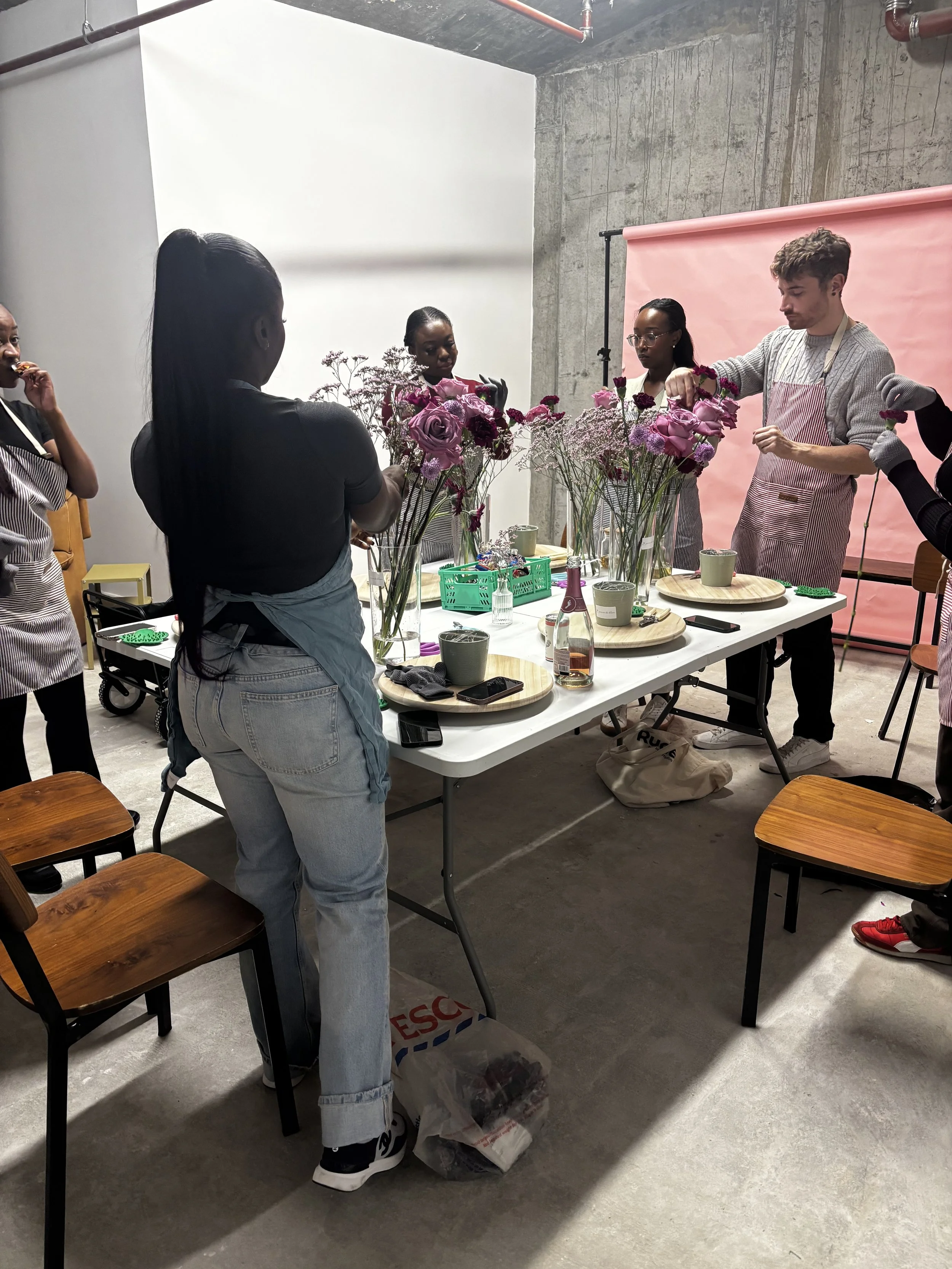 A group of six people decorating with flowers in a studio with a concrete wall and pink backdrop.