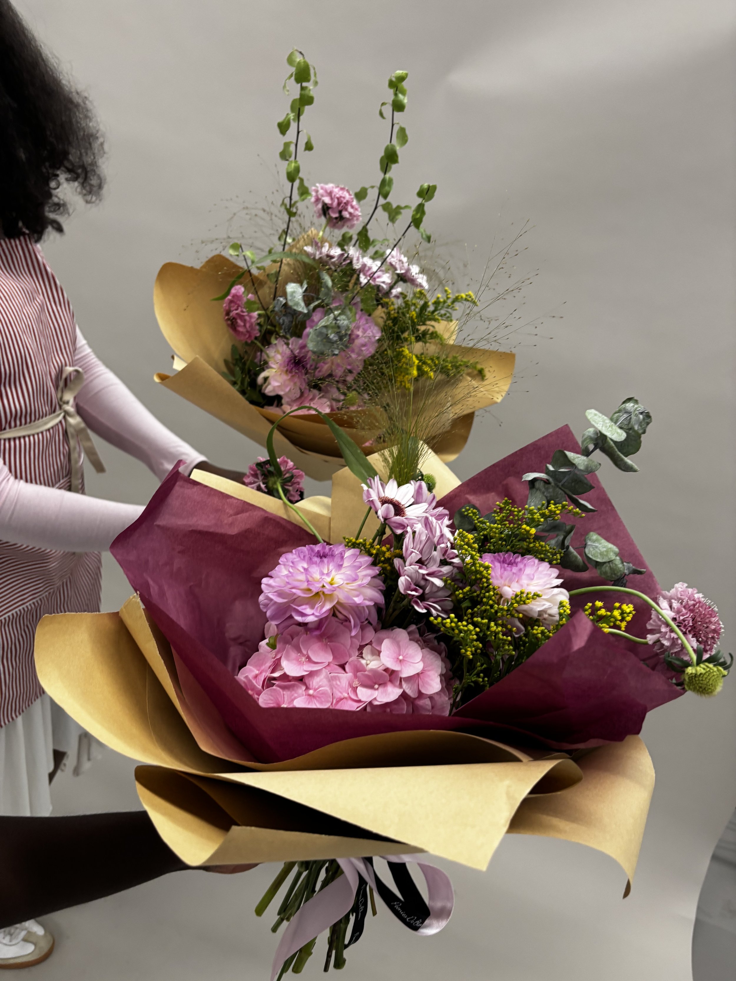 Two bouquets of pink and purple flowers wrapped in brown and pink paper being held by two people.