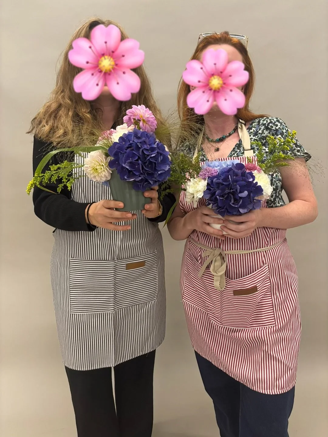 Two women with face obscured by pink flowers holding colorful flower arrangements, wearing striped aprons, standing against a plain beige wall.