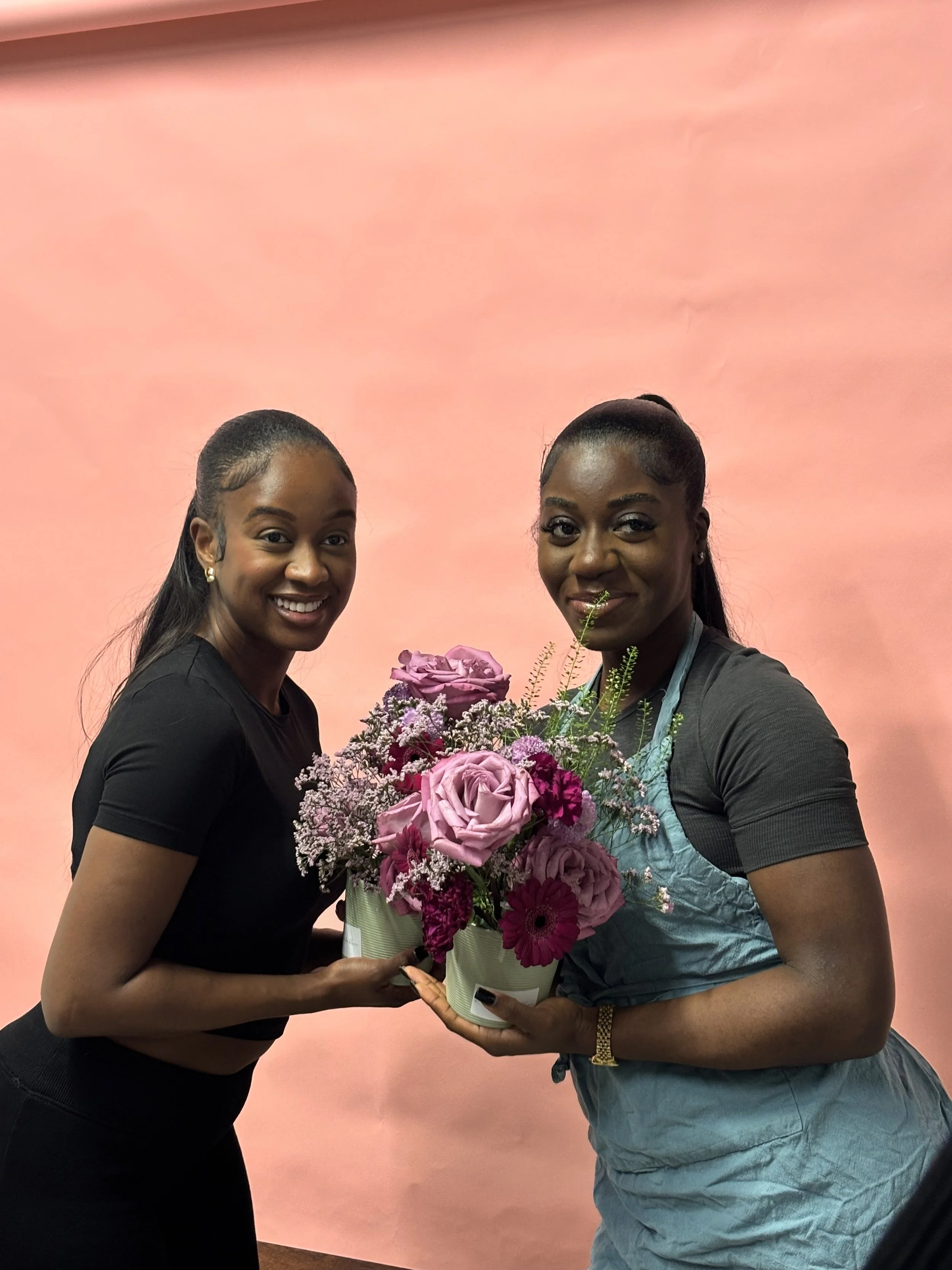 Two women are smiling and holding a pink and purple flower bouquet in front of a pink background.