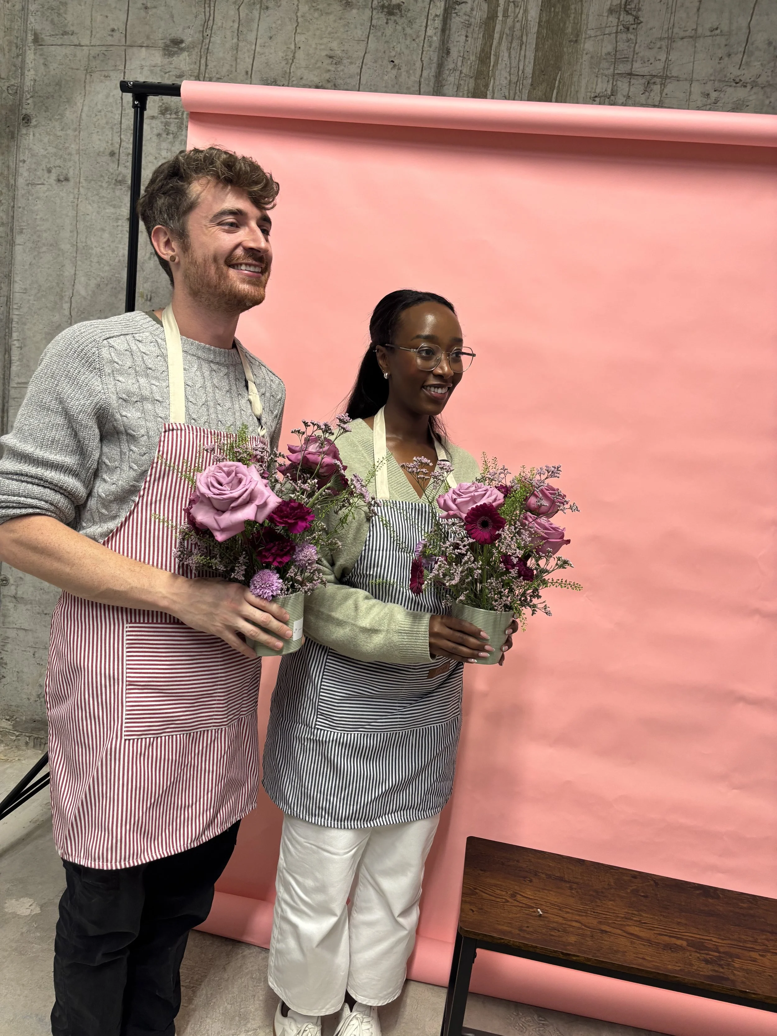 Two people holding flower arrangements in front of a pink backdrop, wearing striped aprons, smiling.