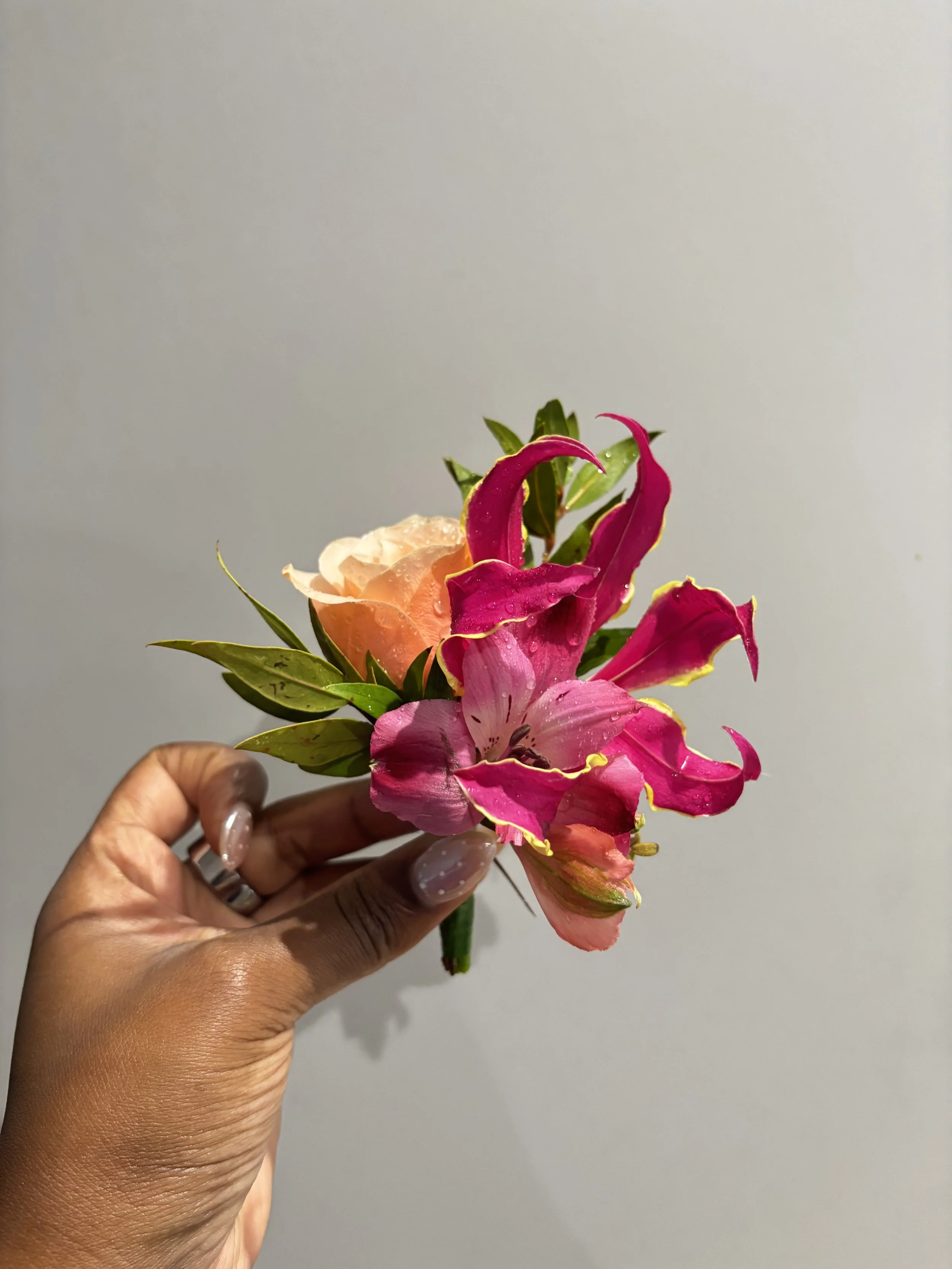 A hand holding a small bouquet of colorful flowers including pink, peach, and purple blooms with green leaves against a plain background.
