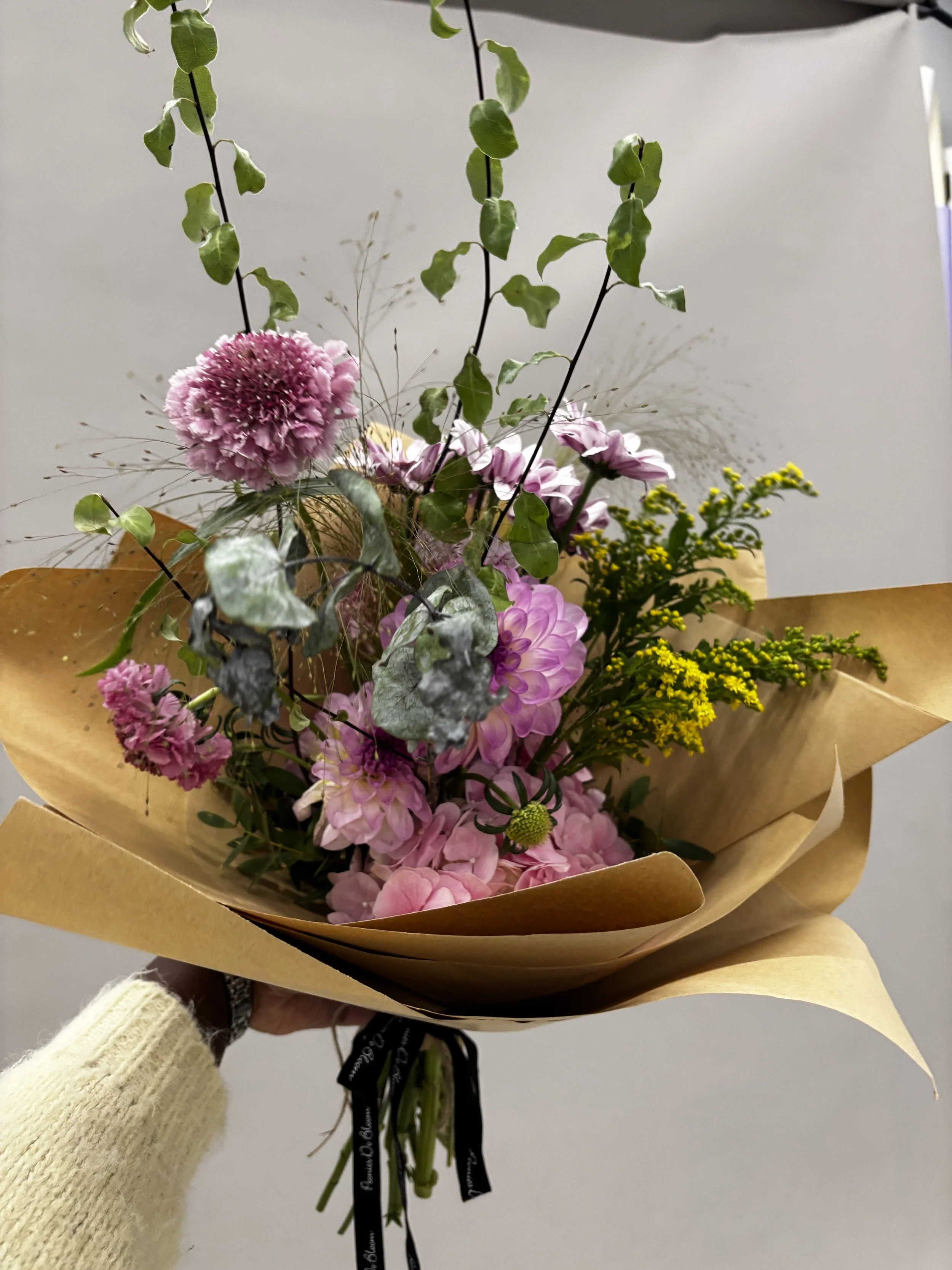 A hand holding a bouquet of pink, purple, and yellow flowers wrapped in brown paper.